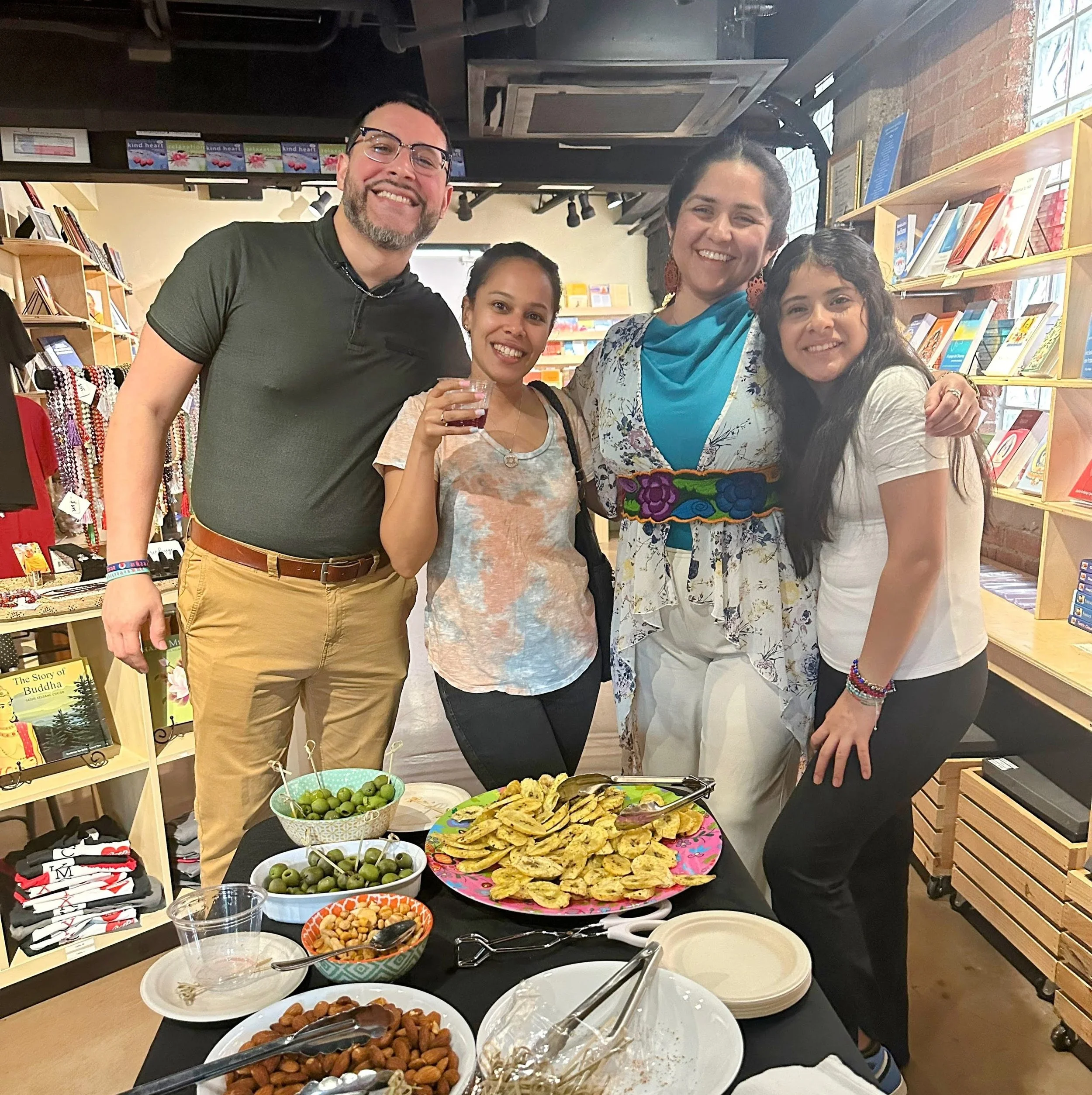 Four people smiling and posing together at a gathering with food on a table, inside a bookstore or café. The table has plates of chips, nuts, olives, and a drink. The background shows bookshelves and store displays.