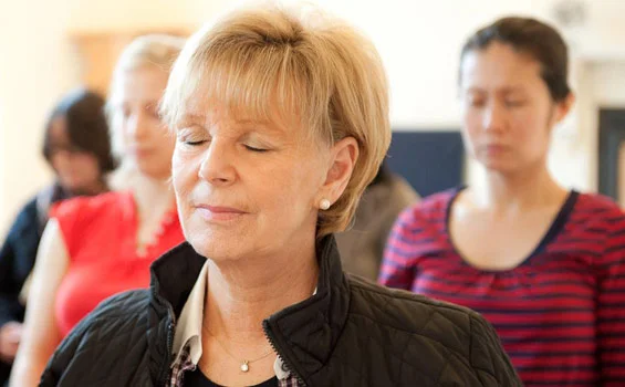 A group of women, including an older woman with short blonde hair and a black jacket, are sitting and appear to be meditating or with eyes closed in a peaceful state.