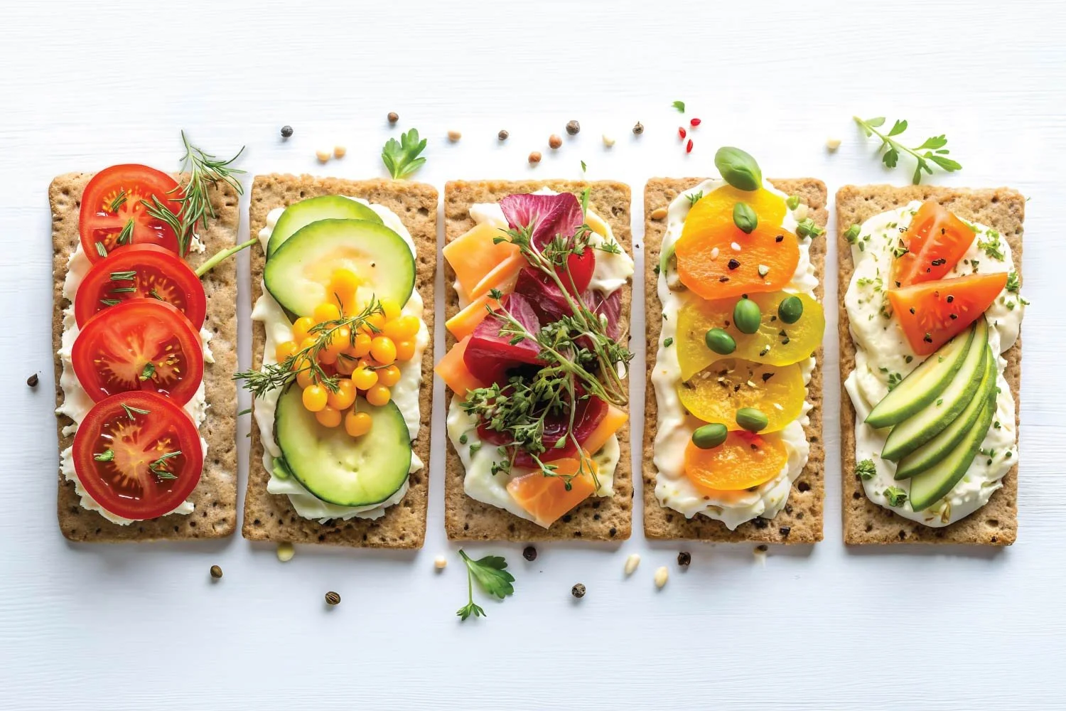 A row of five cream cheese crispbreads topped with assorted fresh vegetables, herbs, and smoked salmon on a white background.