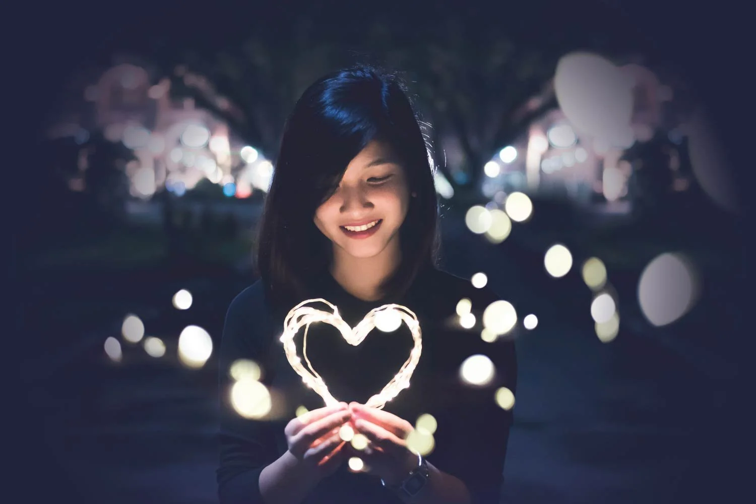 A smiling woman looks down at a glowing heart made of string lights cupped in her hands, surrounded by soft, sparkling lights against a dark night background.