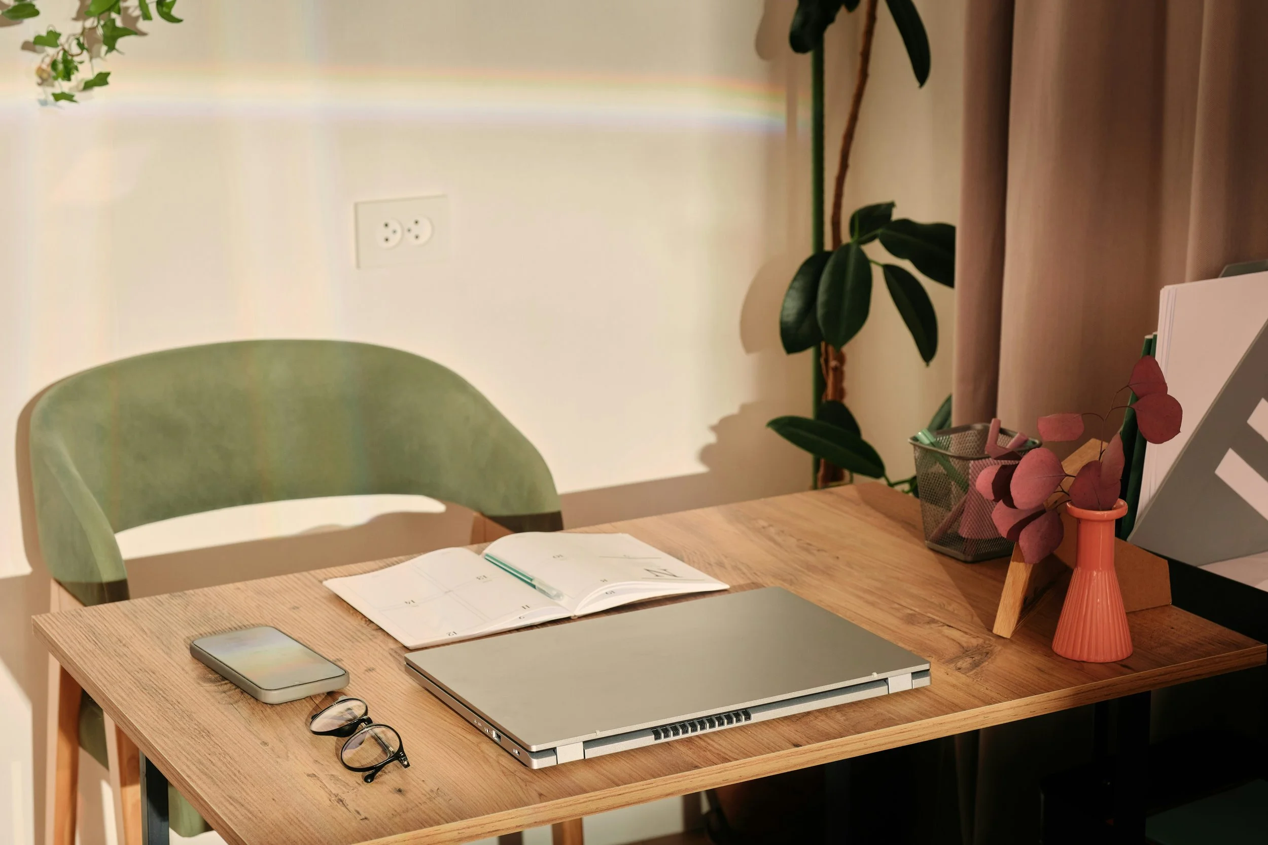 A wooden desk with a closed laptop, open planner with a pen, smartphone, and glasses on it. There is a green chair, a potted plant with large leaves, a pink vase with dried pink leaves, a container holding pens and pencils, and a TV stand with a laptop and a document holder. The background shows a white wall with a power outlet and beige curtains.