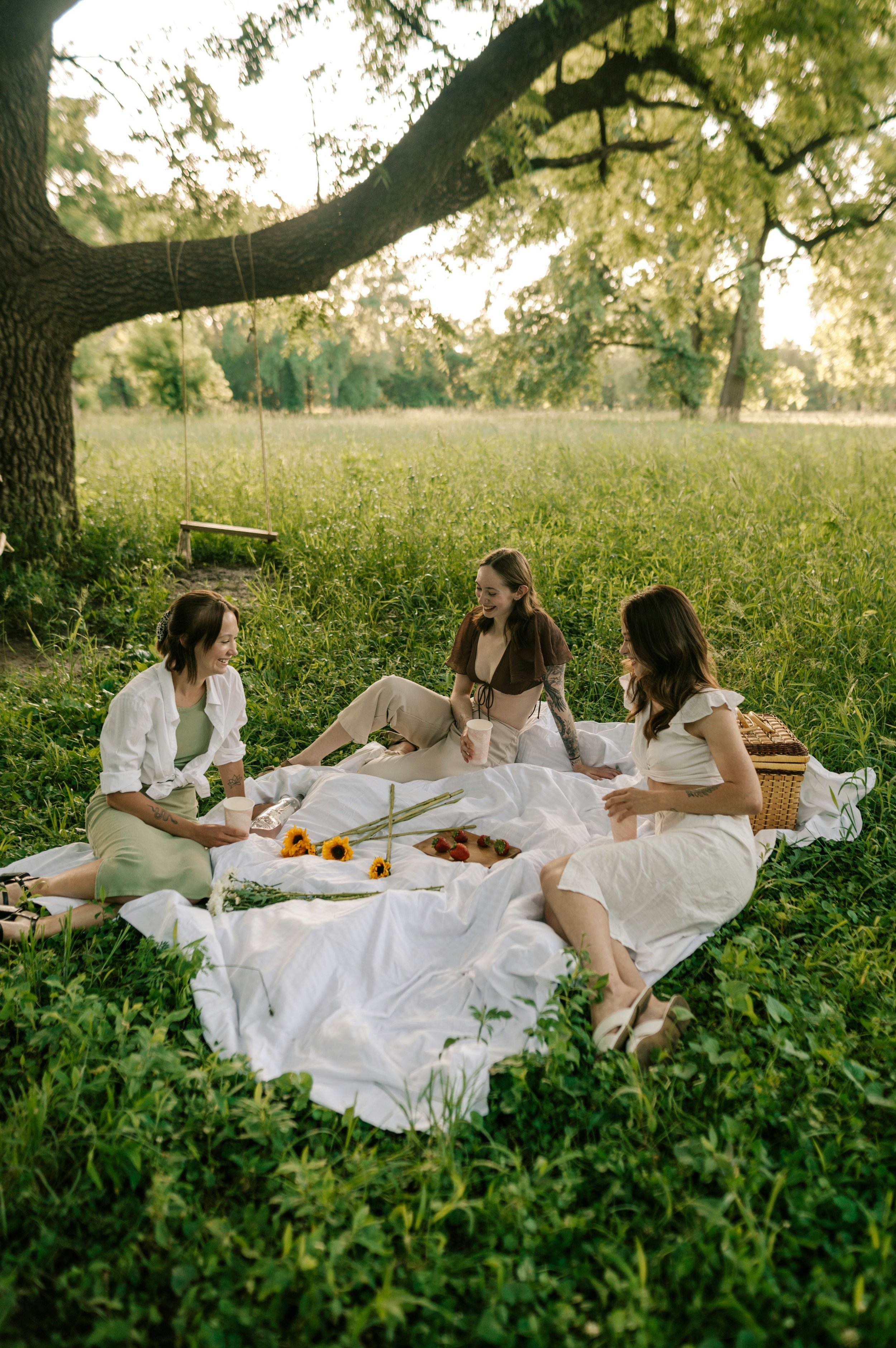Three women having a picnic in a grassy field under a large tree, with a picnic blanket, food, and drinks.