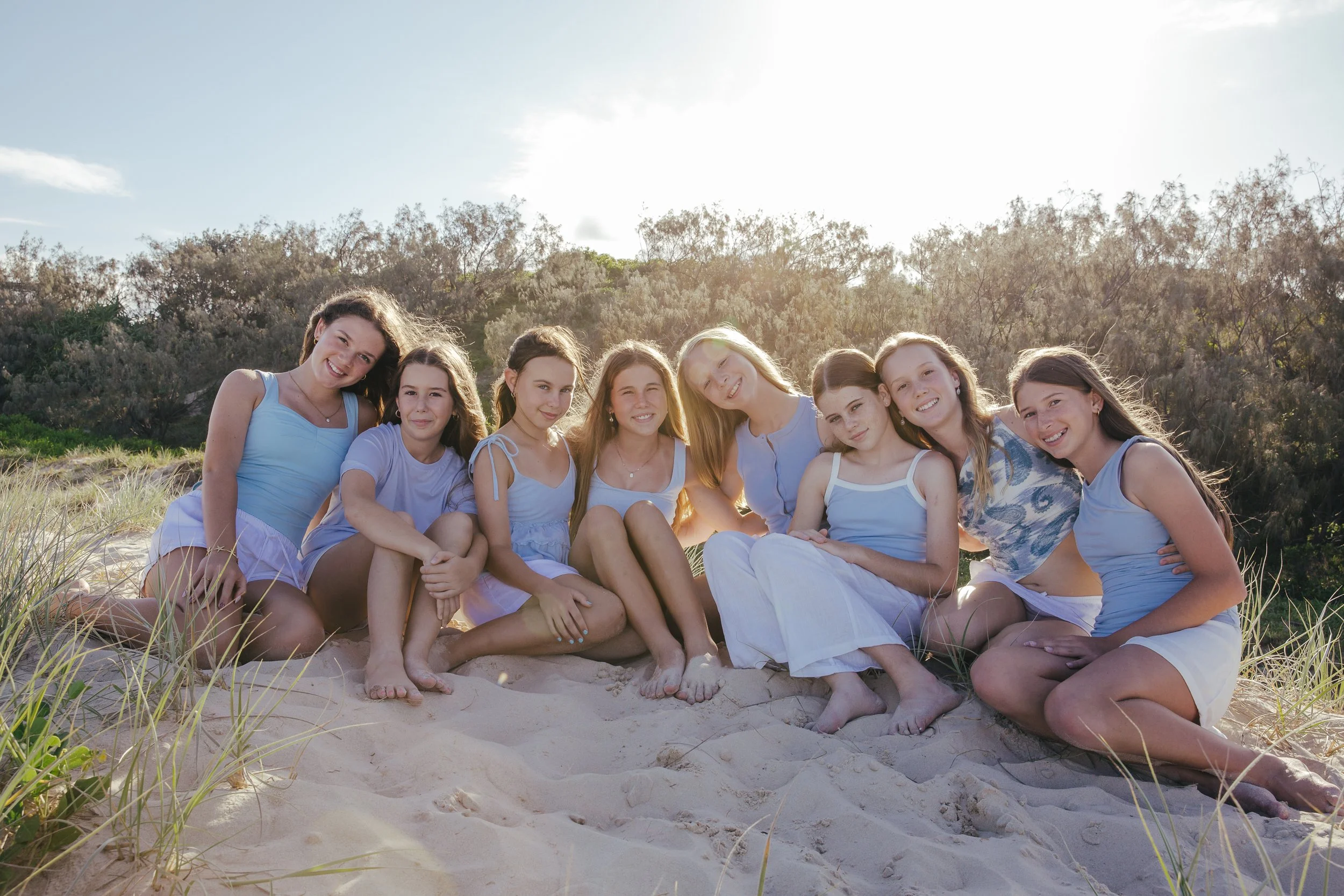 Group of nine girls sitting on the sand at the beach, smiling, wearing casual summer clothes, with trees and sky in the background.