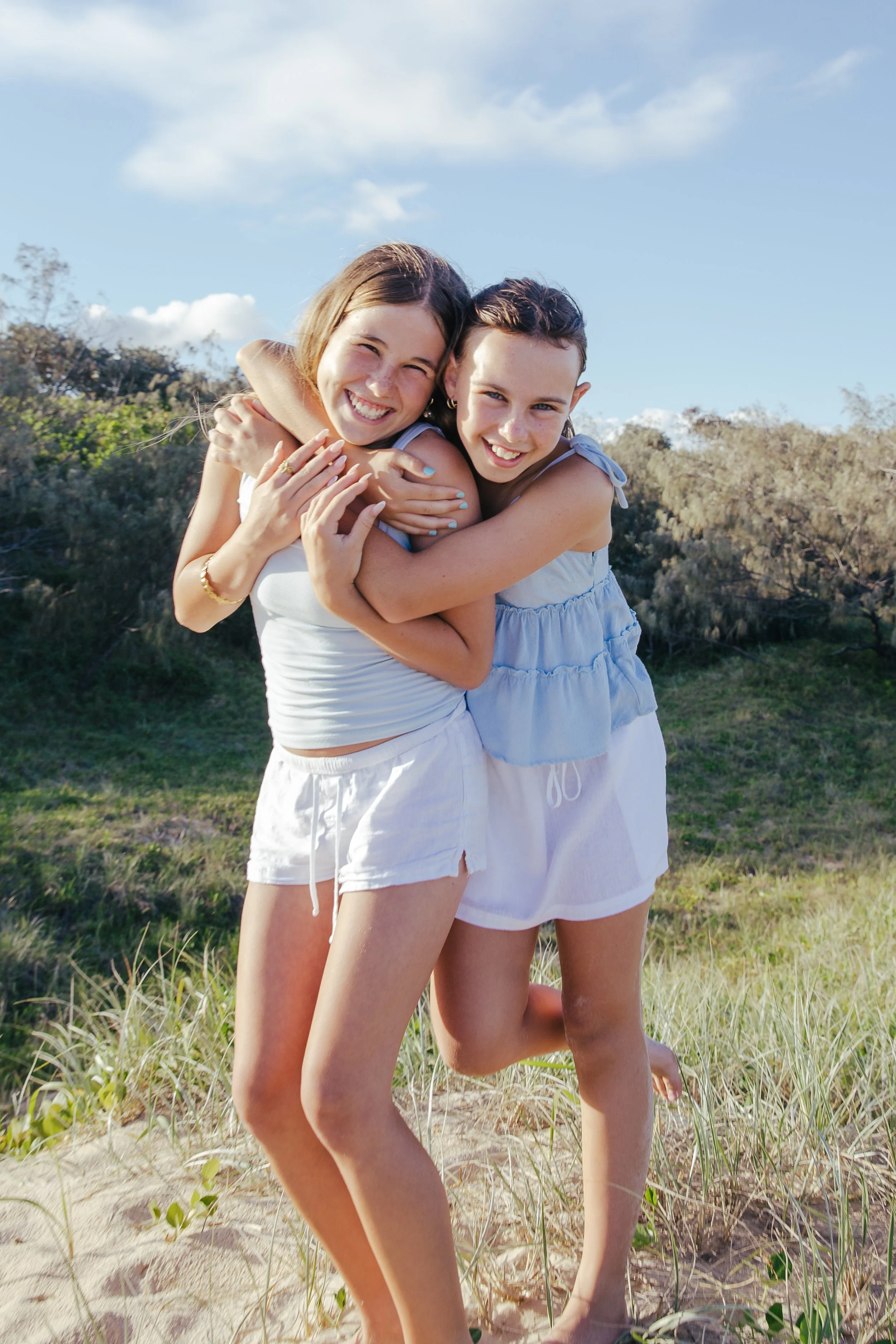 Two young women hugging and smiling outdoors on a sunny day, standing on grassy sand dunes with trees and blue sky in the background.
