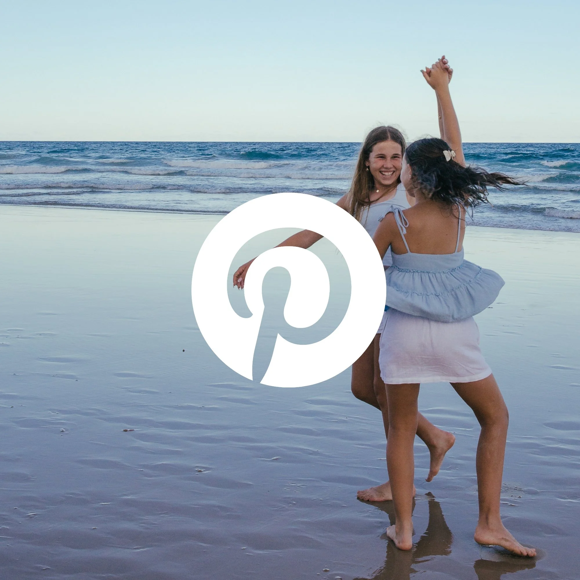 Two women standing in shallow ocean water, smiling, with one raising her arm in the air during sunset or sunrise with a clear sky.