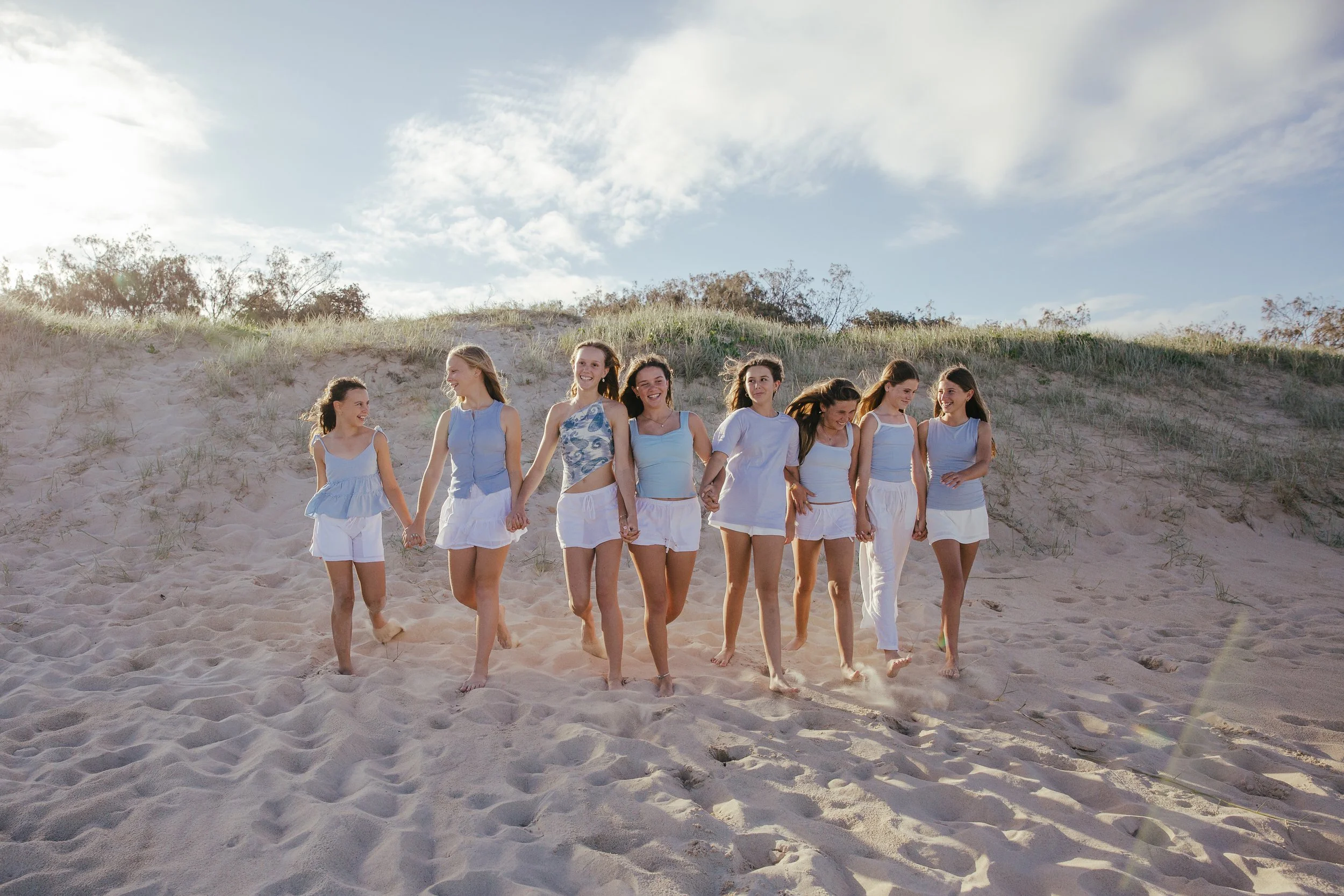 Eight women walking hand-in-hand on a sandy beach with dunes and grass in the background, dressed in light blue and white summer outfits.