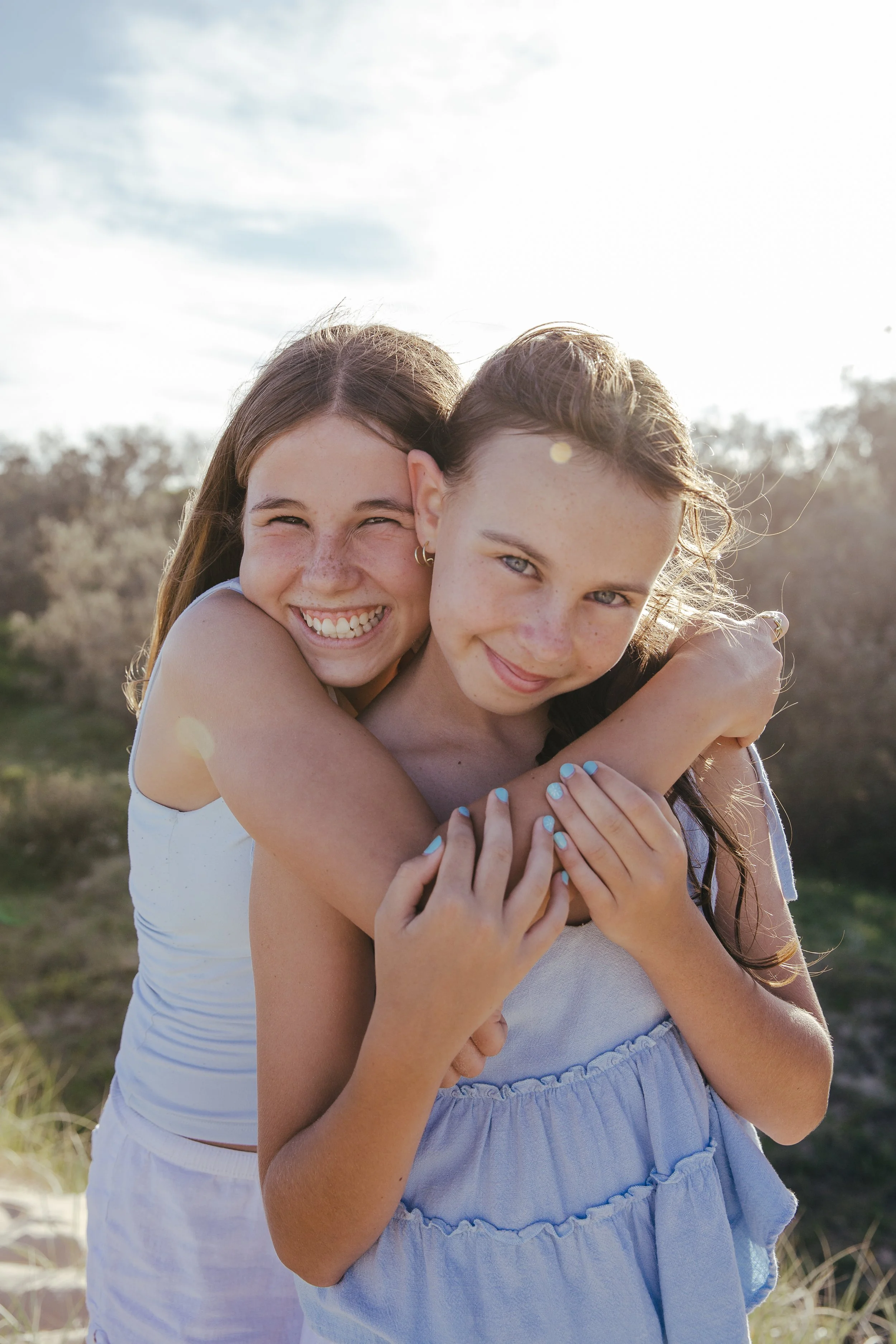 Two girls happily hugging outdoors with nature in the background, smiling at the camera, bathed in sunlight.