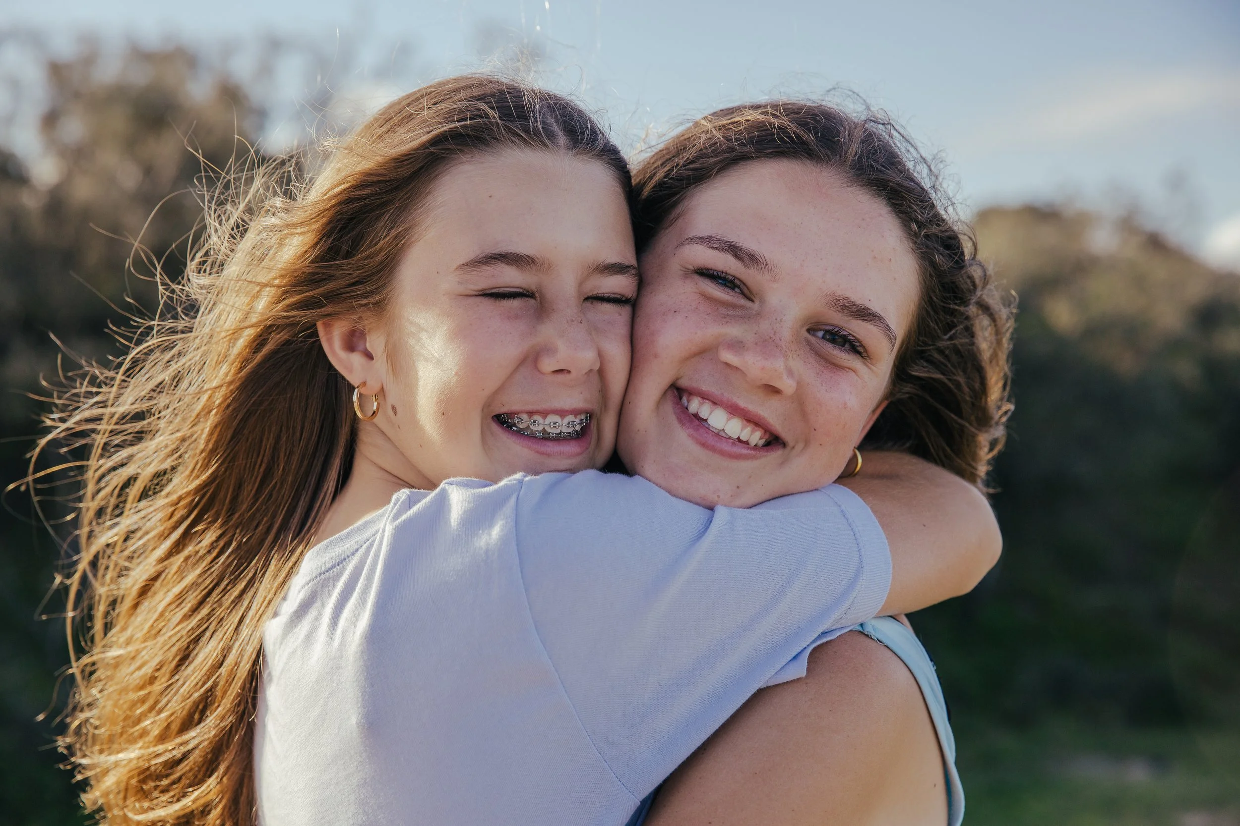 Two young women hugging outdoors, smiling joyfully, with one girl having braces and both with long hair, in a natural setting with trees and blue sky.