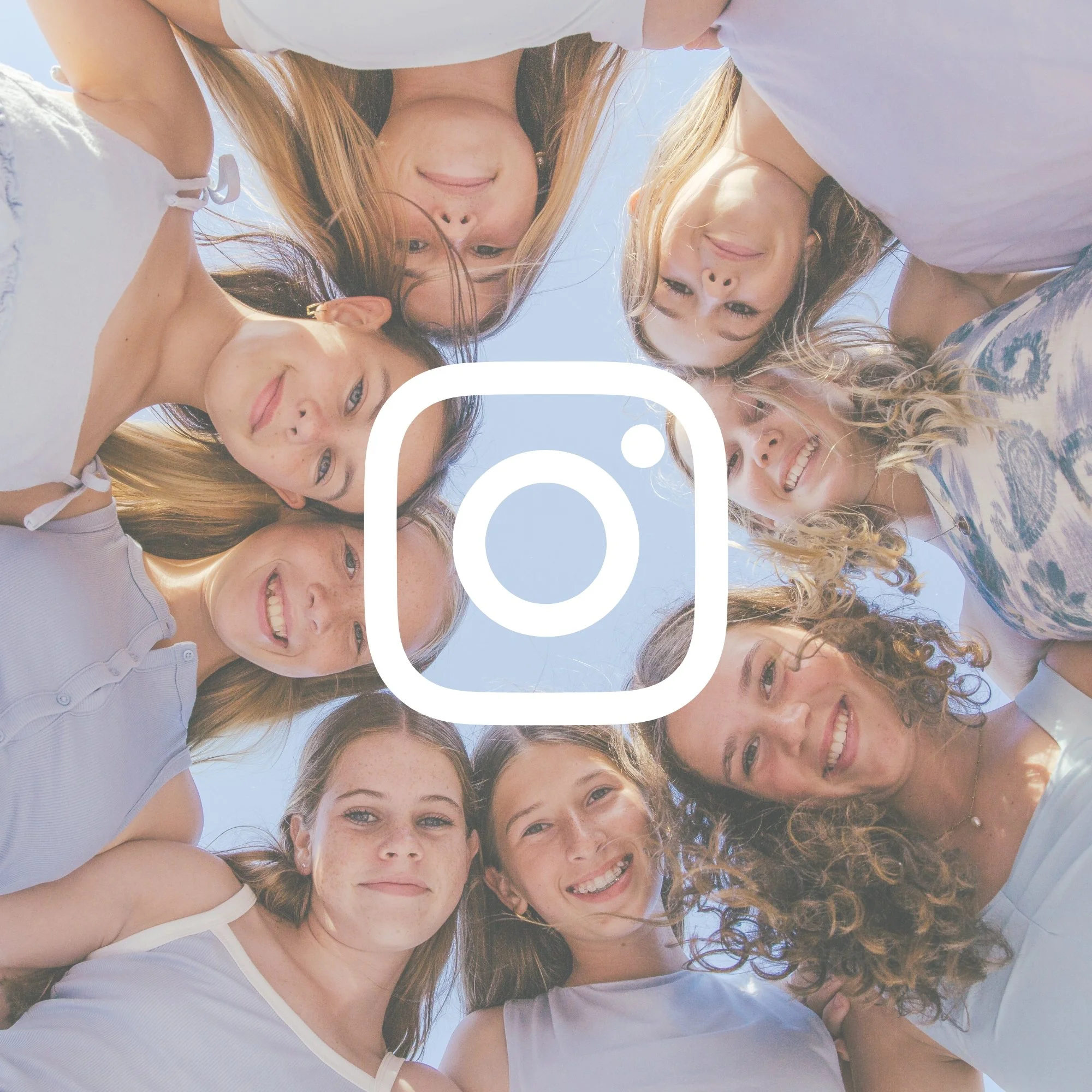 A group of women and a man looking down into the camera, forming a circle with their heads outdoors on a sunny day.