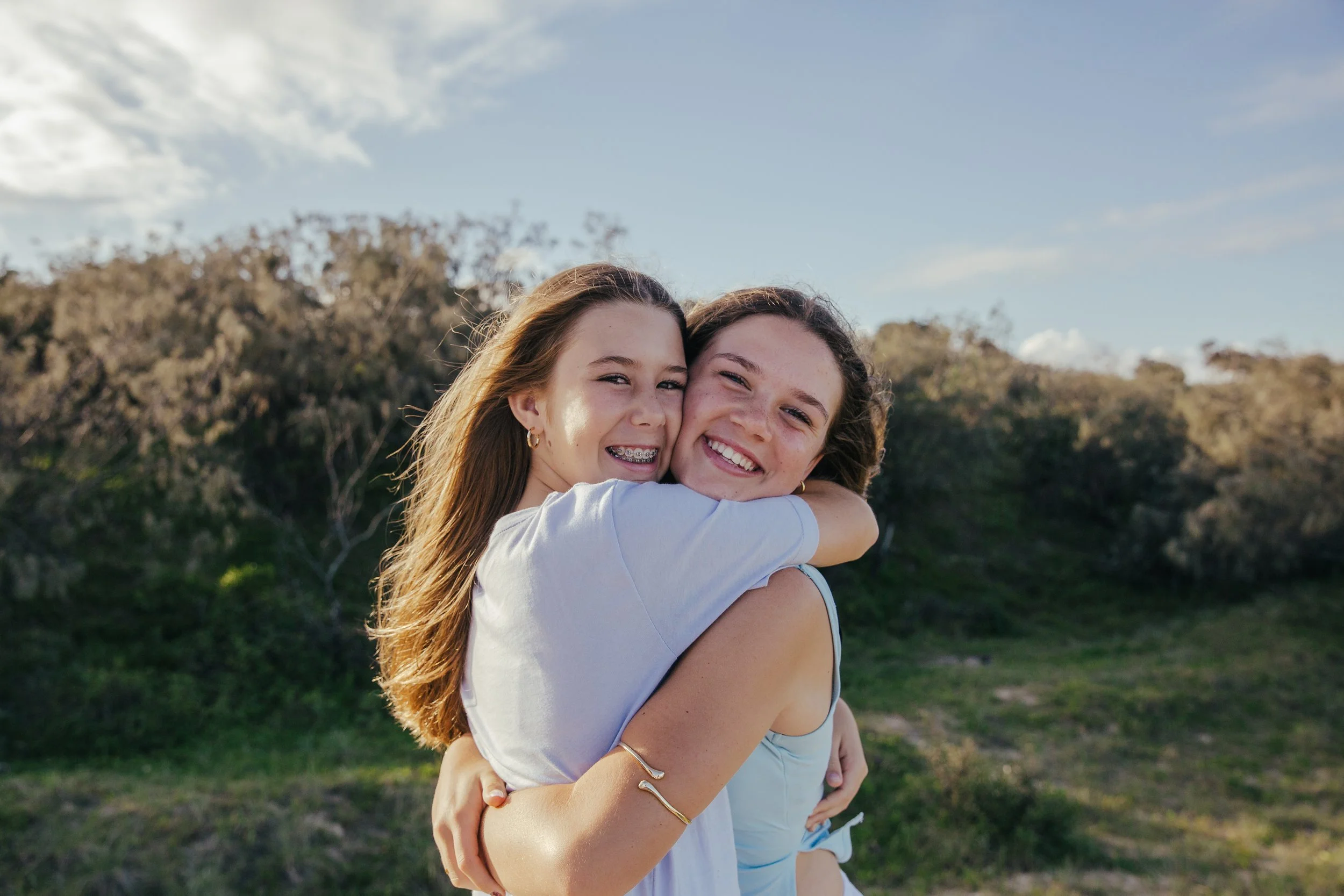 Two young girls hugging outdoors on a sunny day, with blue sky and trees in the background.