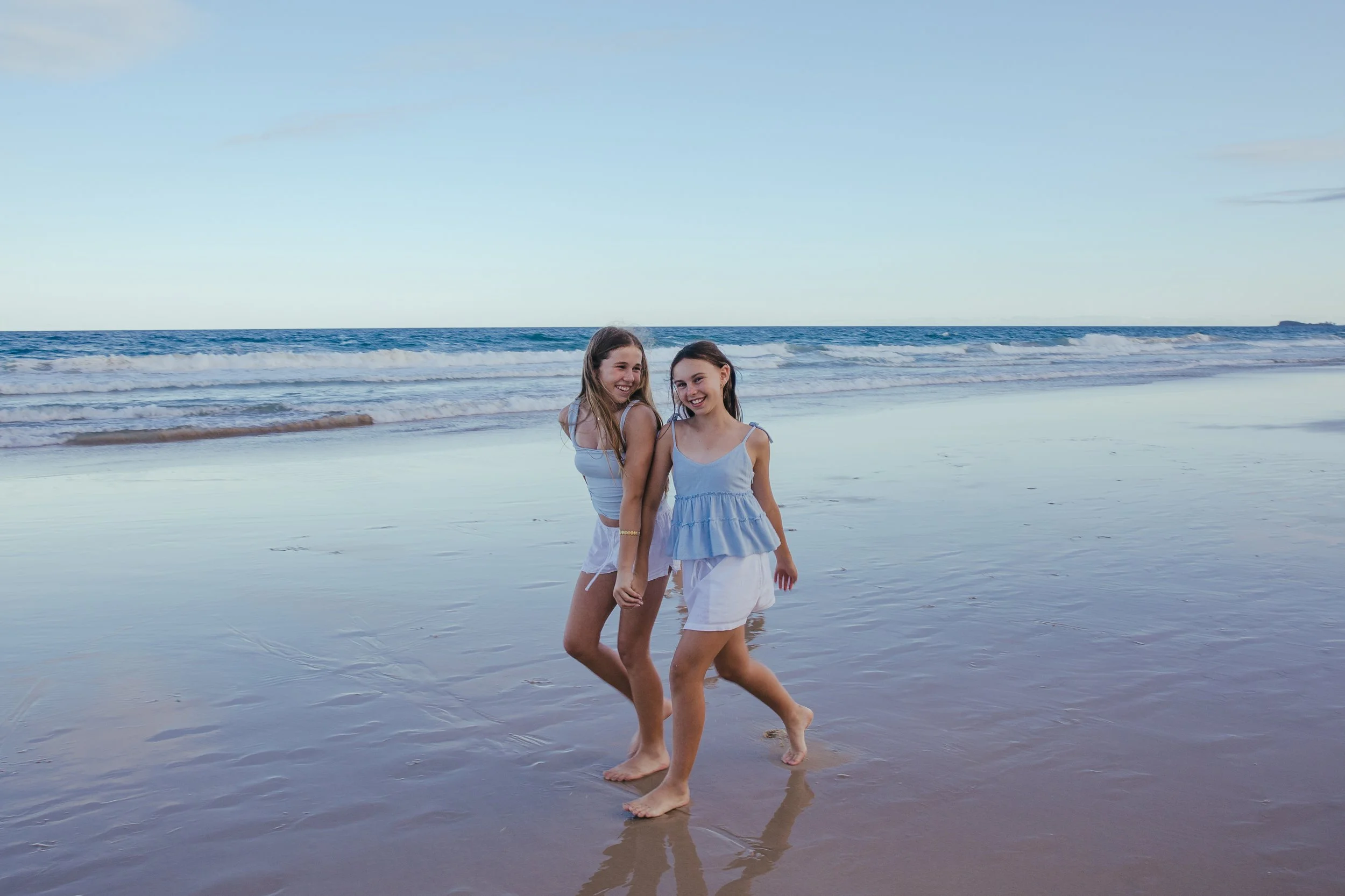 Two young girls smiling and holding hands while walking barefoot on wet sand at the beach with gentle waves in the background during daytime.
