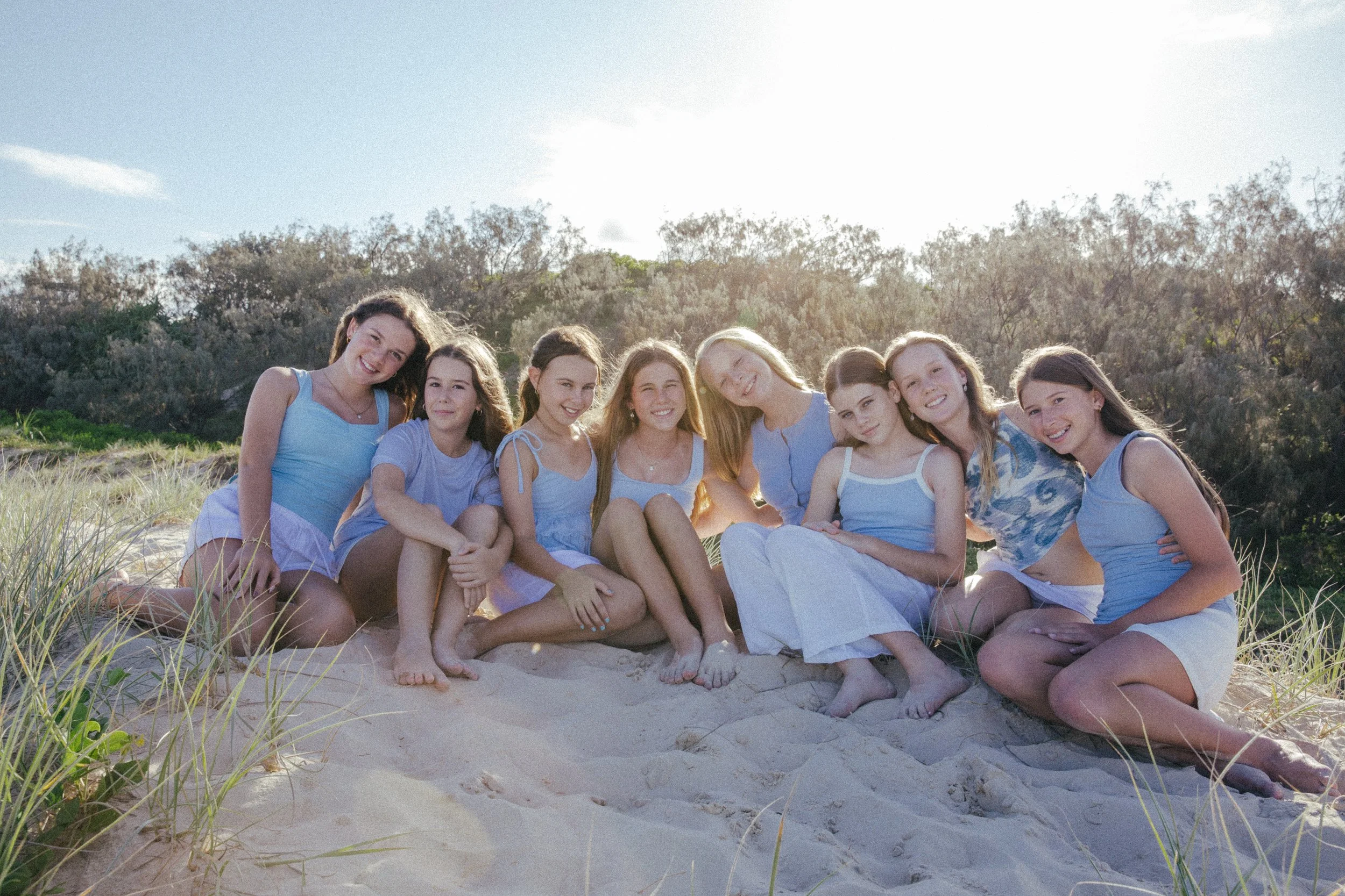 Group of nine girls sitting on the sand at the beach, smiling and enjoying the sunlight, with trees and blue sky in the background.