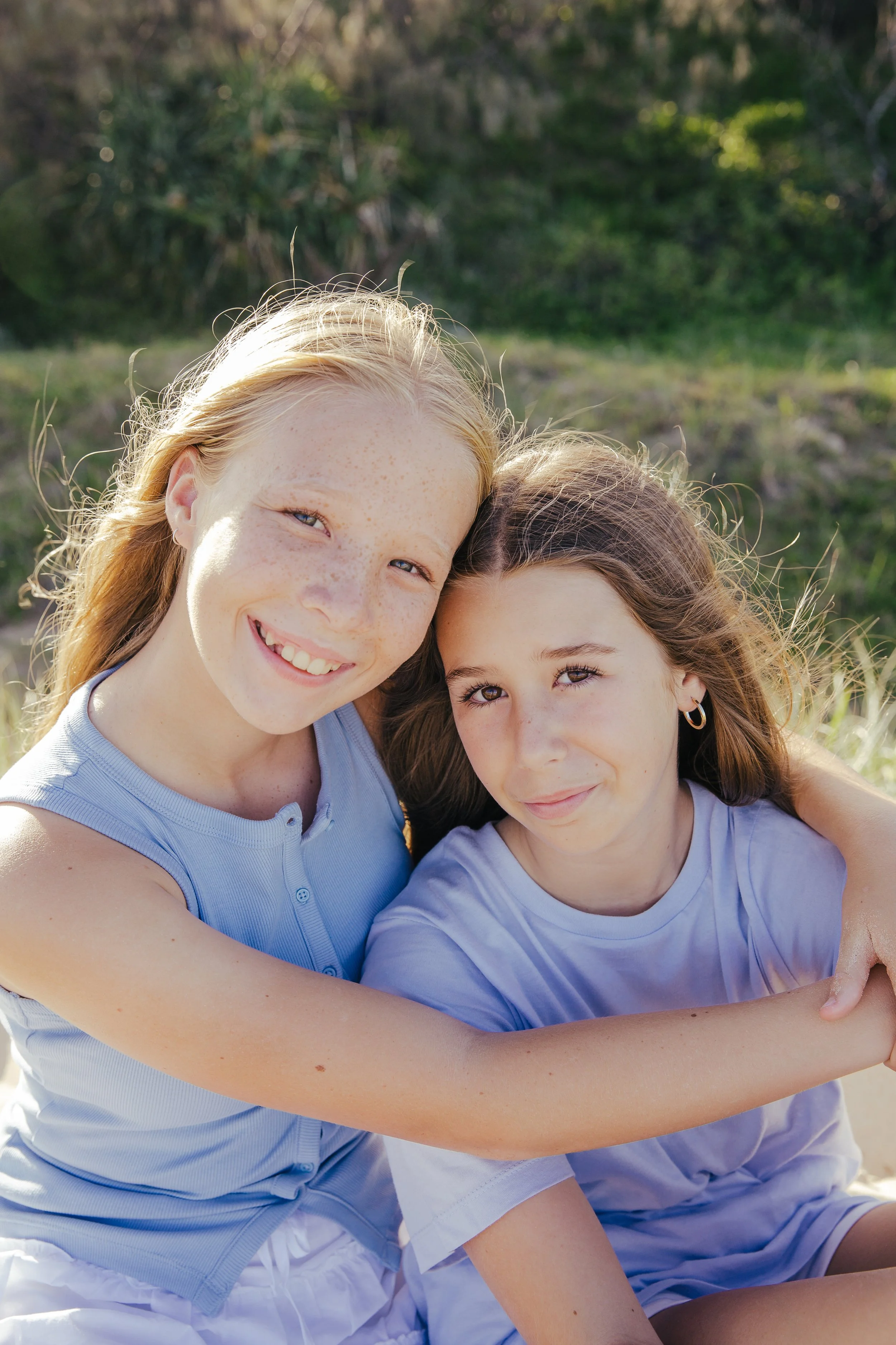 Two young girls sitting outdoors, smiling and hugging each other on a sunny day.