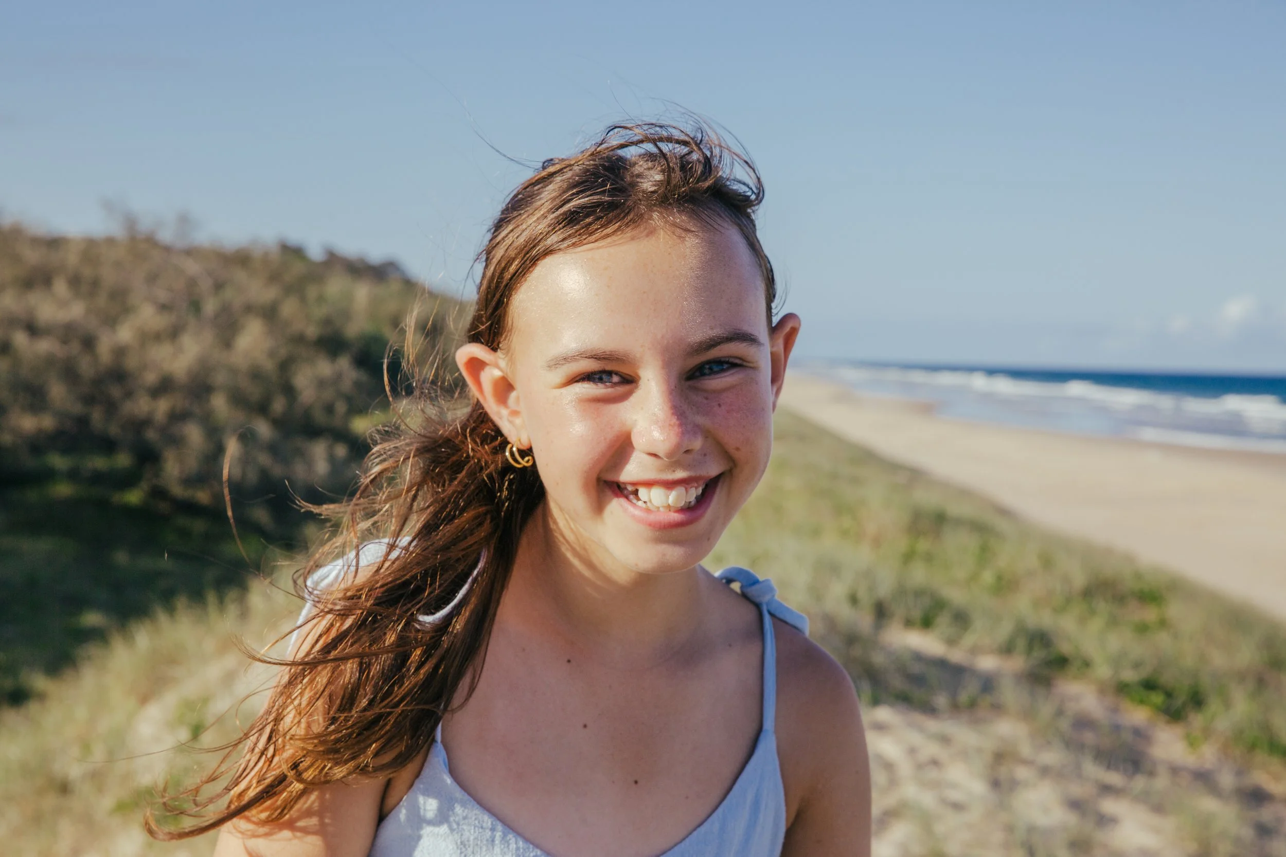 A young girl with long brown hair smiling on a beach with sand dunes and the ocean in the background.