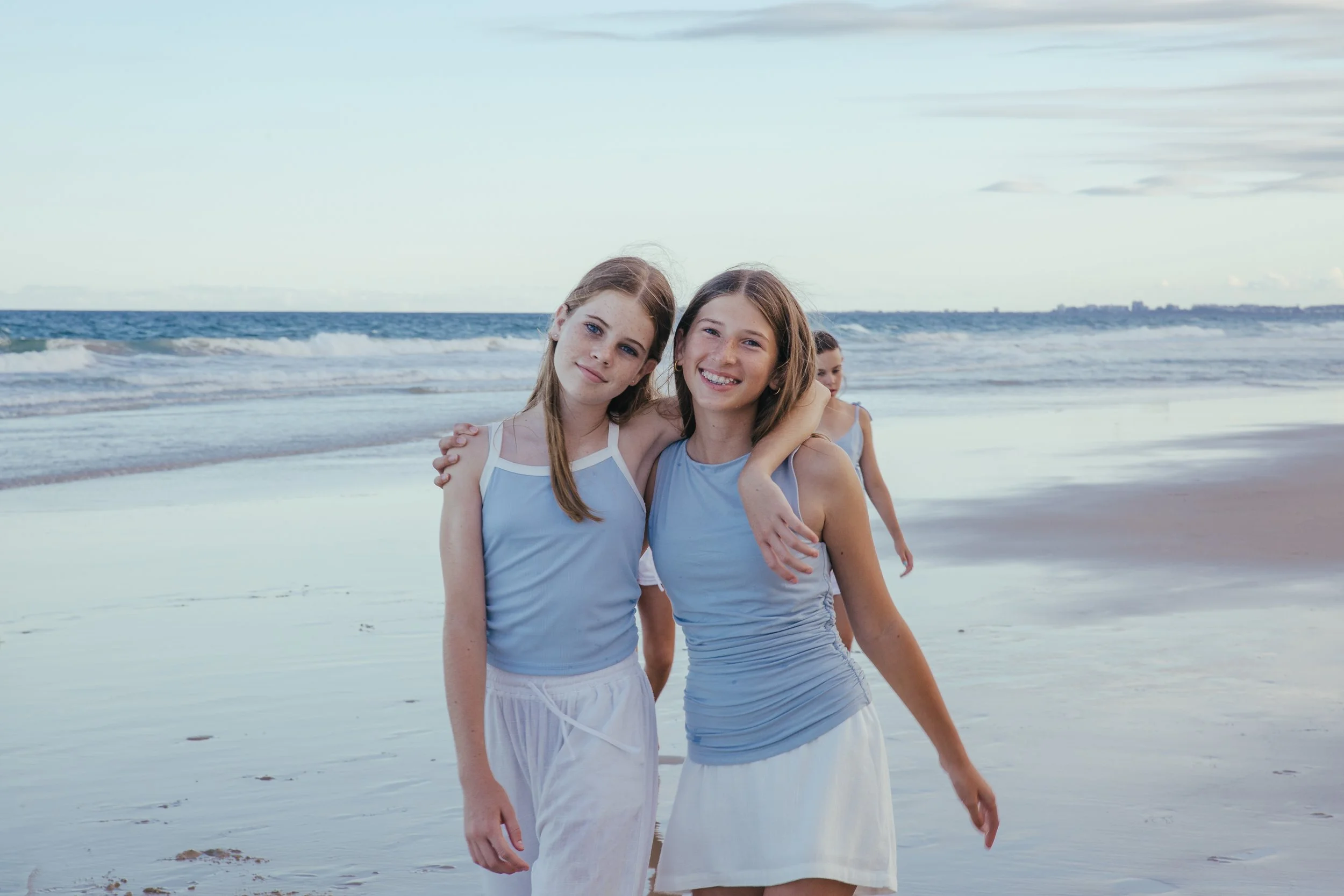 Two young girls in matching blue tank tops and white skirts standing on a sandy beach with the ocean waves in the background, smiling with their arms around each other.