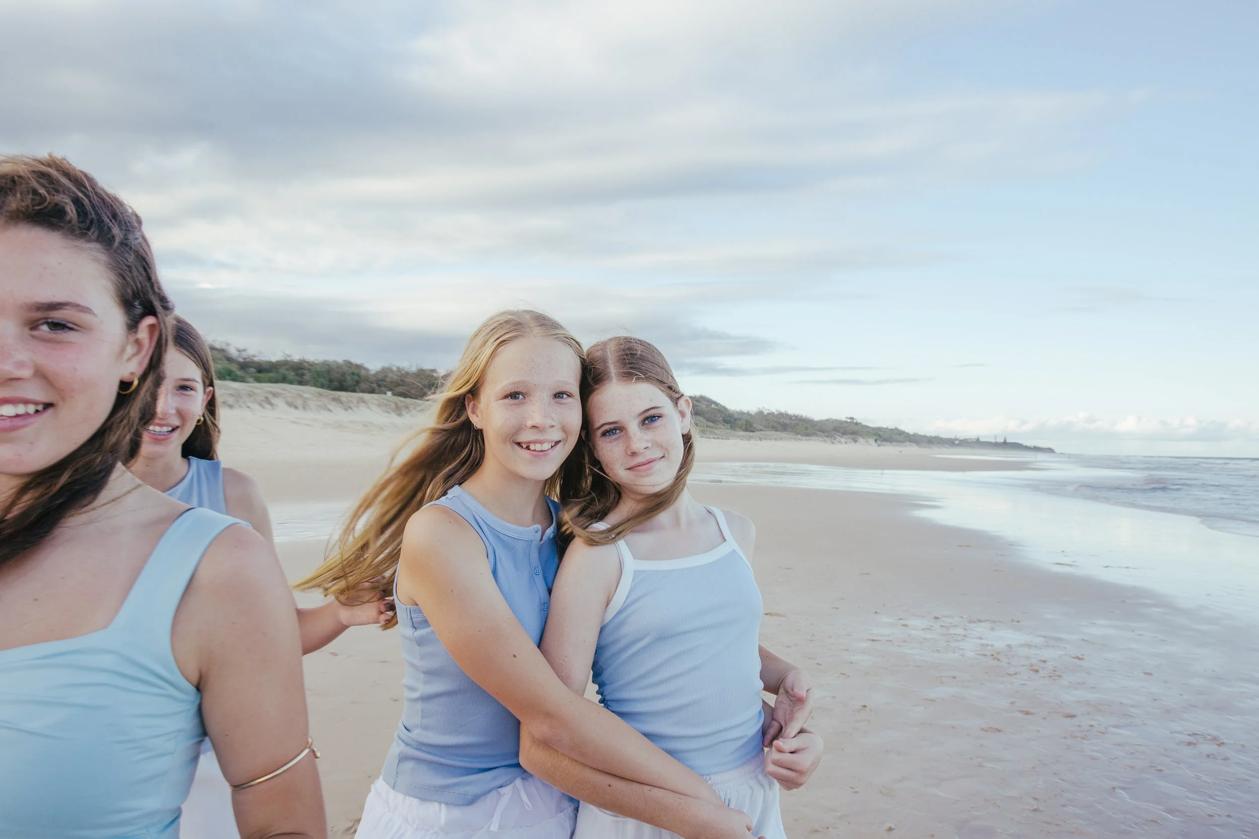 Four young girls at the beach, standing close together, smiling, with sand dunes and ocean in the background under a partly cloudy sky.