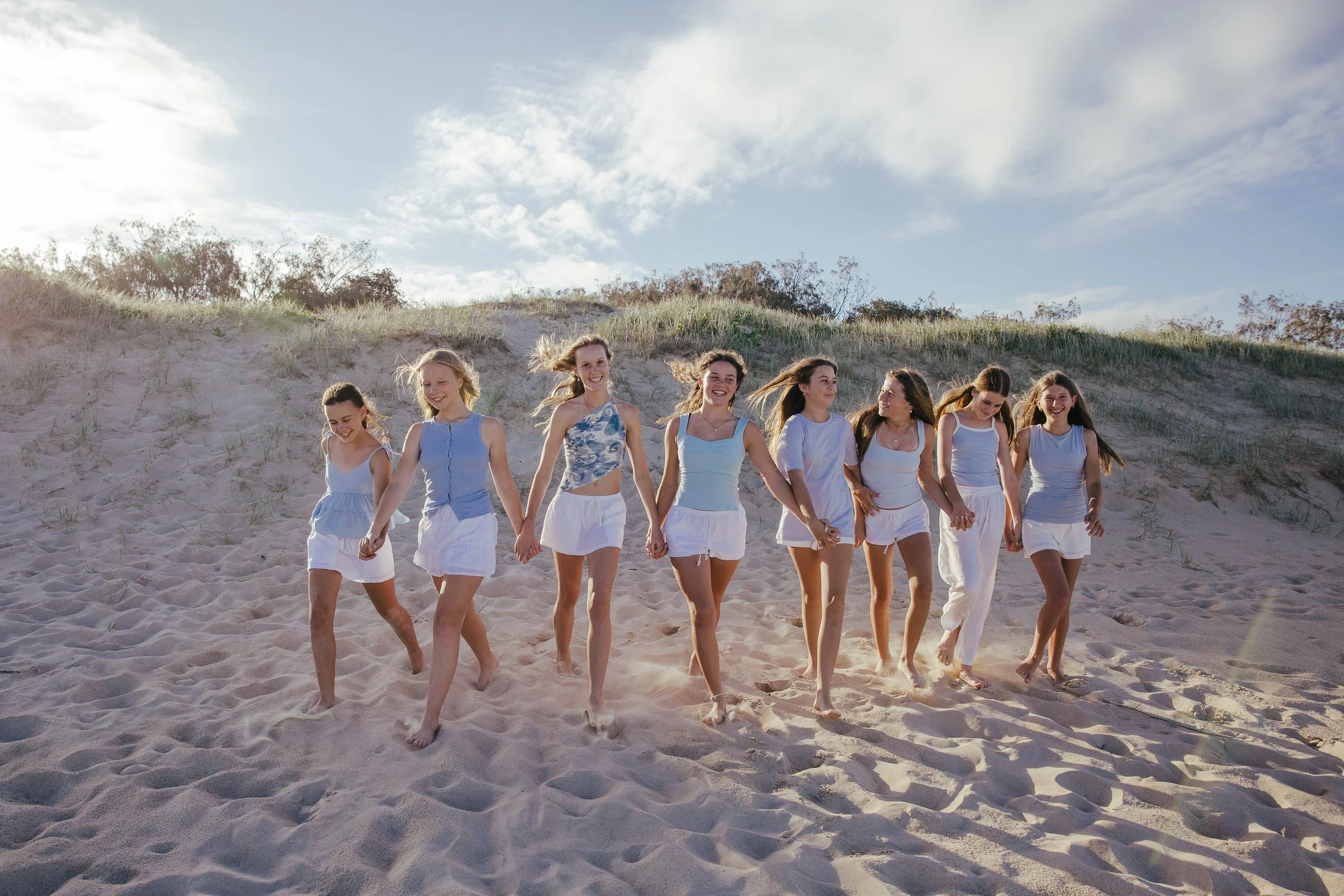 A group of nine young women dressed in white and light blue summer outfits walk hand in hand on a sandy beach with dunes and trees in the background under a partly cloudy sky.
