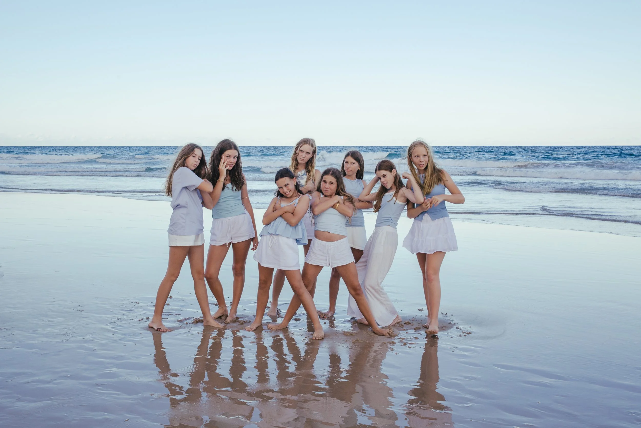 Group of nine young girls standing on the wet sand at the beach, wearing casual summer clothing, with the ocean and blue sky in the background.