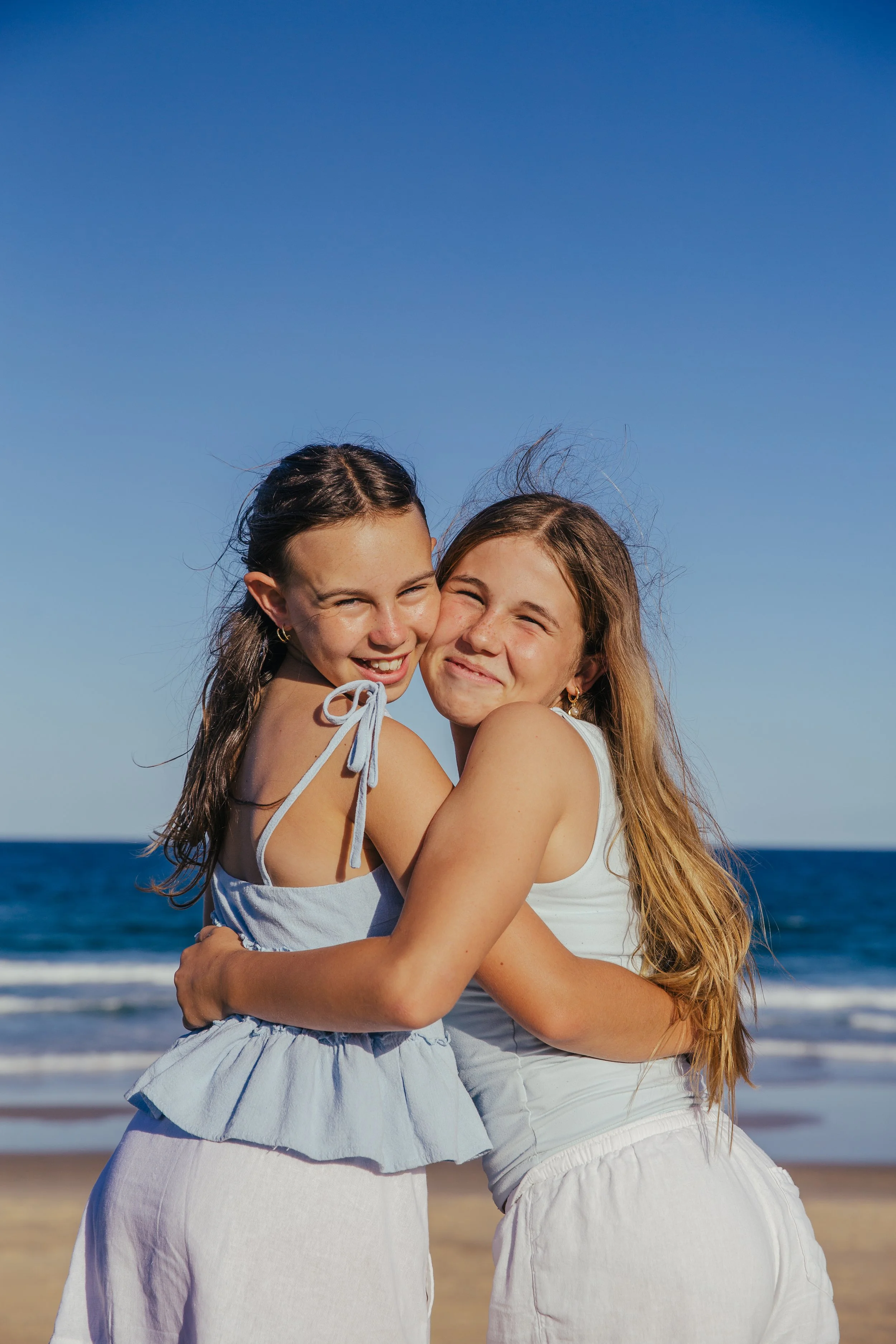 Two young girls hugging on the beach with the ocean and a clear blue sky in the background.