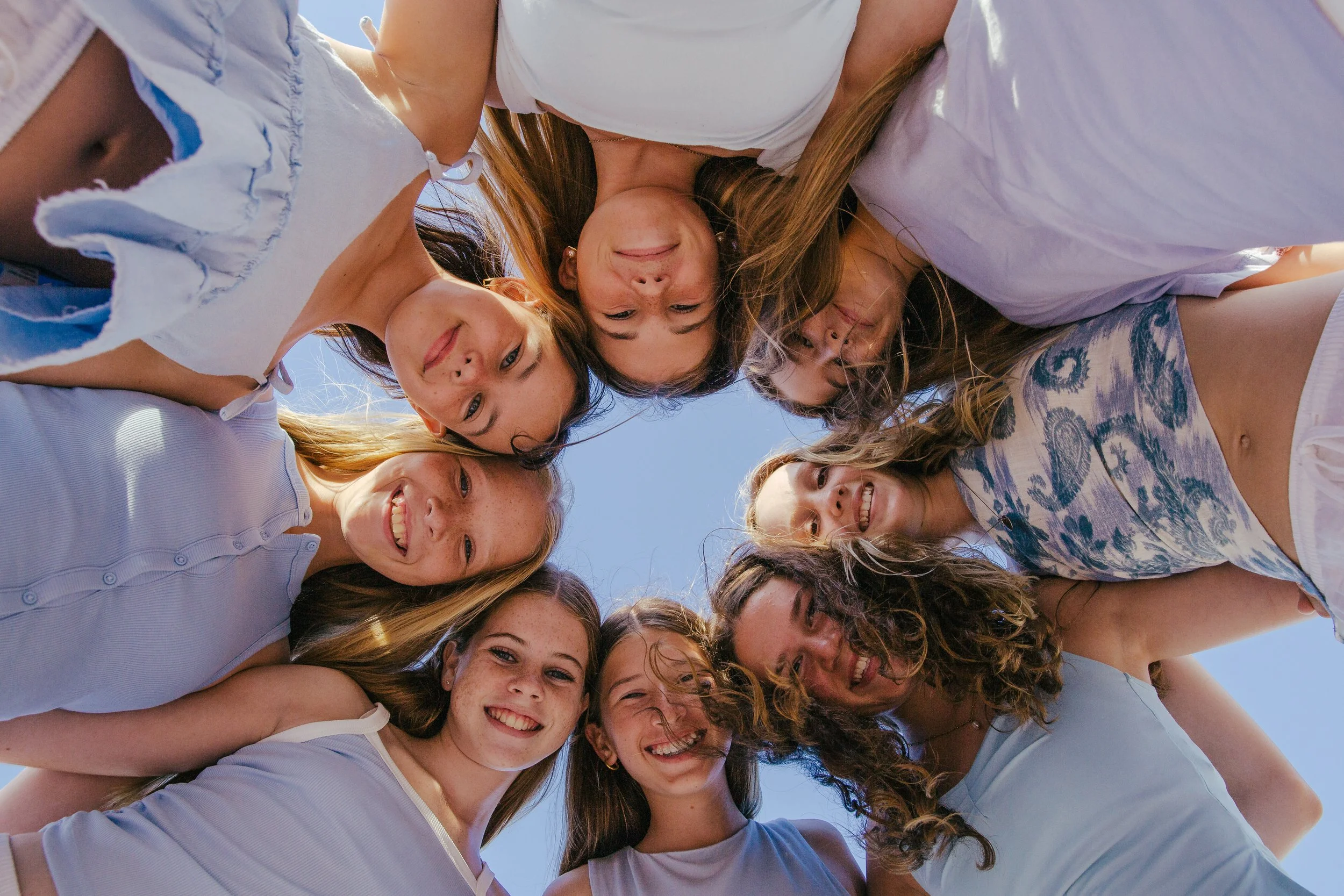 A group of nine young women forming a circle and looking down at the camera, smiling, outdoors under a clear blue sky.