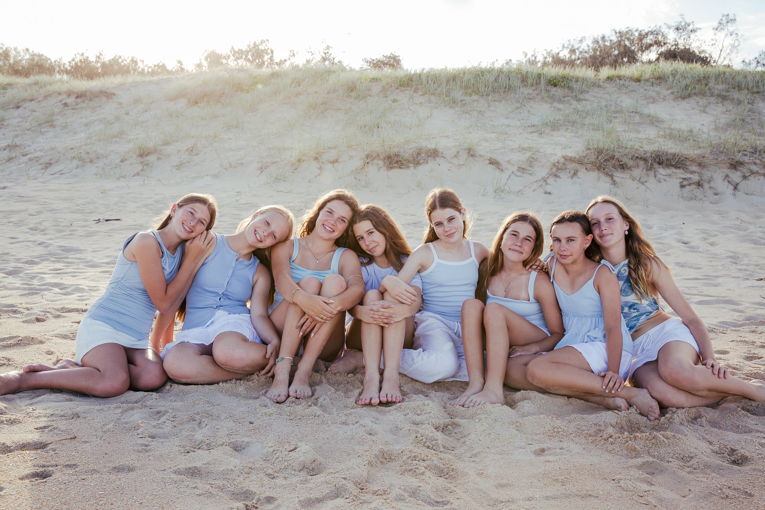 A group of nine young girls sitting on the sandy beach, smiling and relaxing together, with sand dunes and sparse vegetation in the background.
