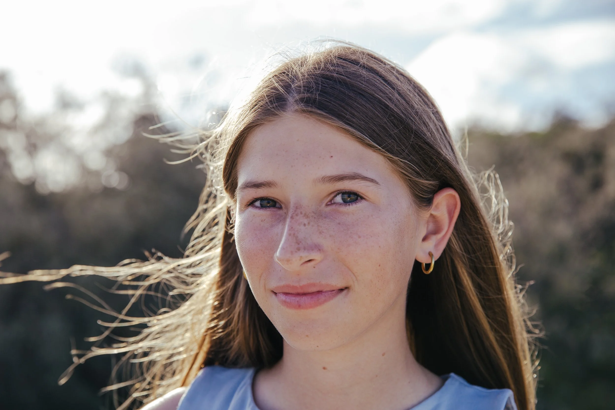 A young girl with long brown hair, blue eyes, and freckles, standing outdoors with sunlight, wearing a sleeveless top and gold hoop earrings.