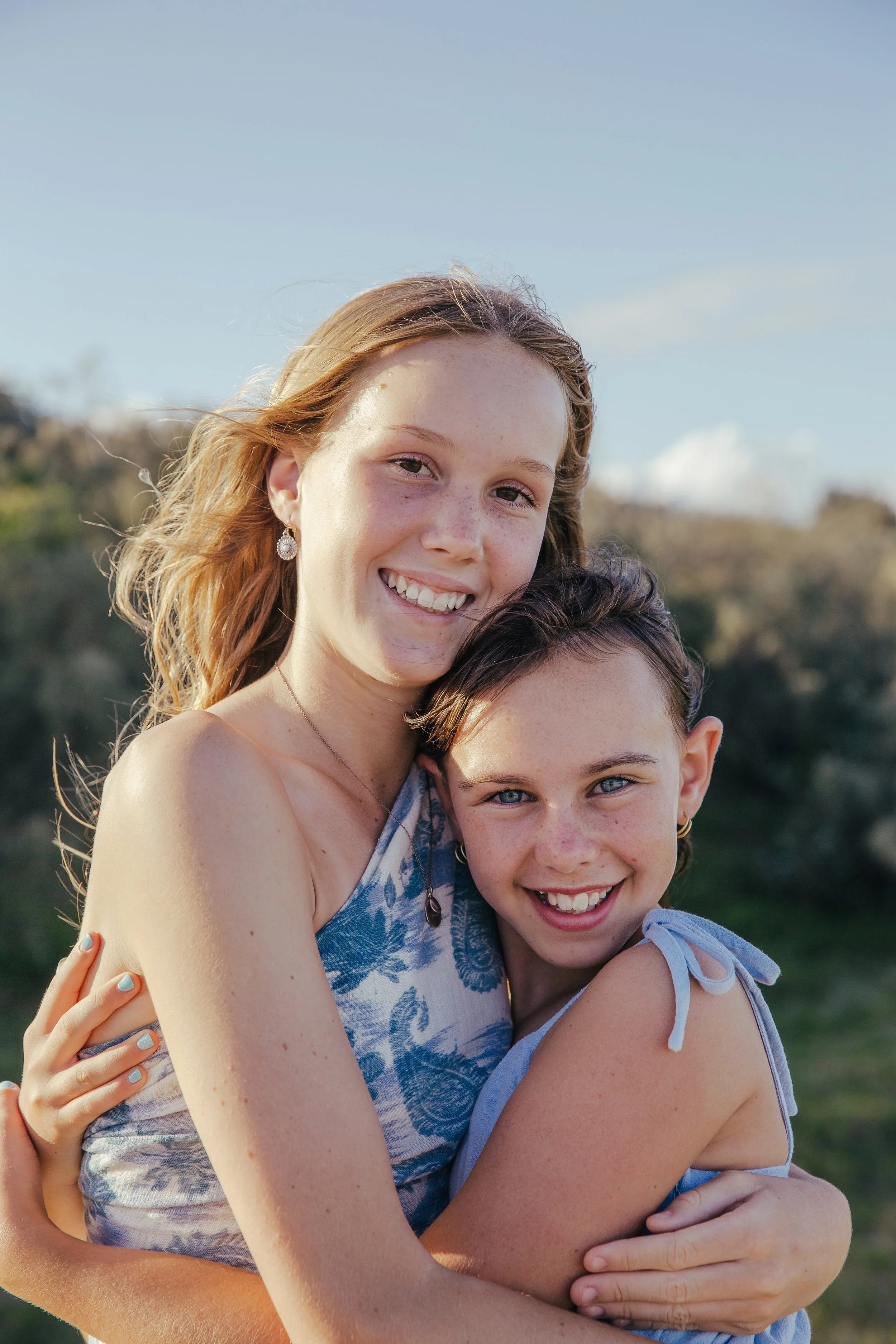 Two girls smiling and hugging outdoors on a sunny day.