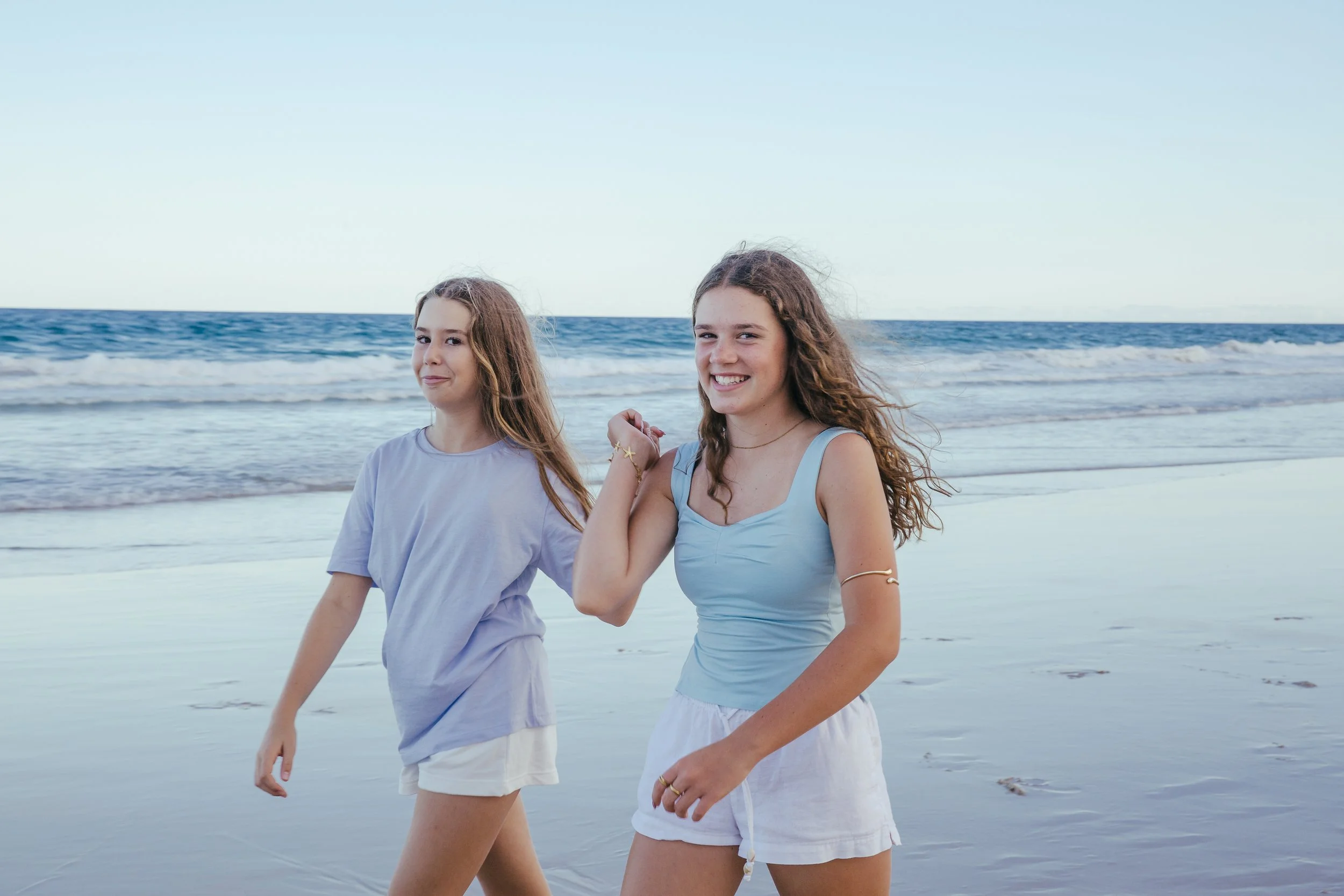 Two young women walking along the beach, holding hands, with the ocean and sky in the background.