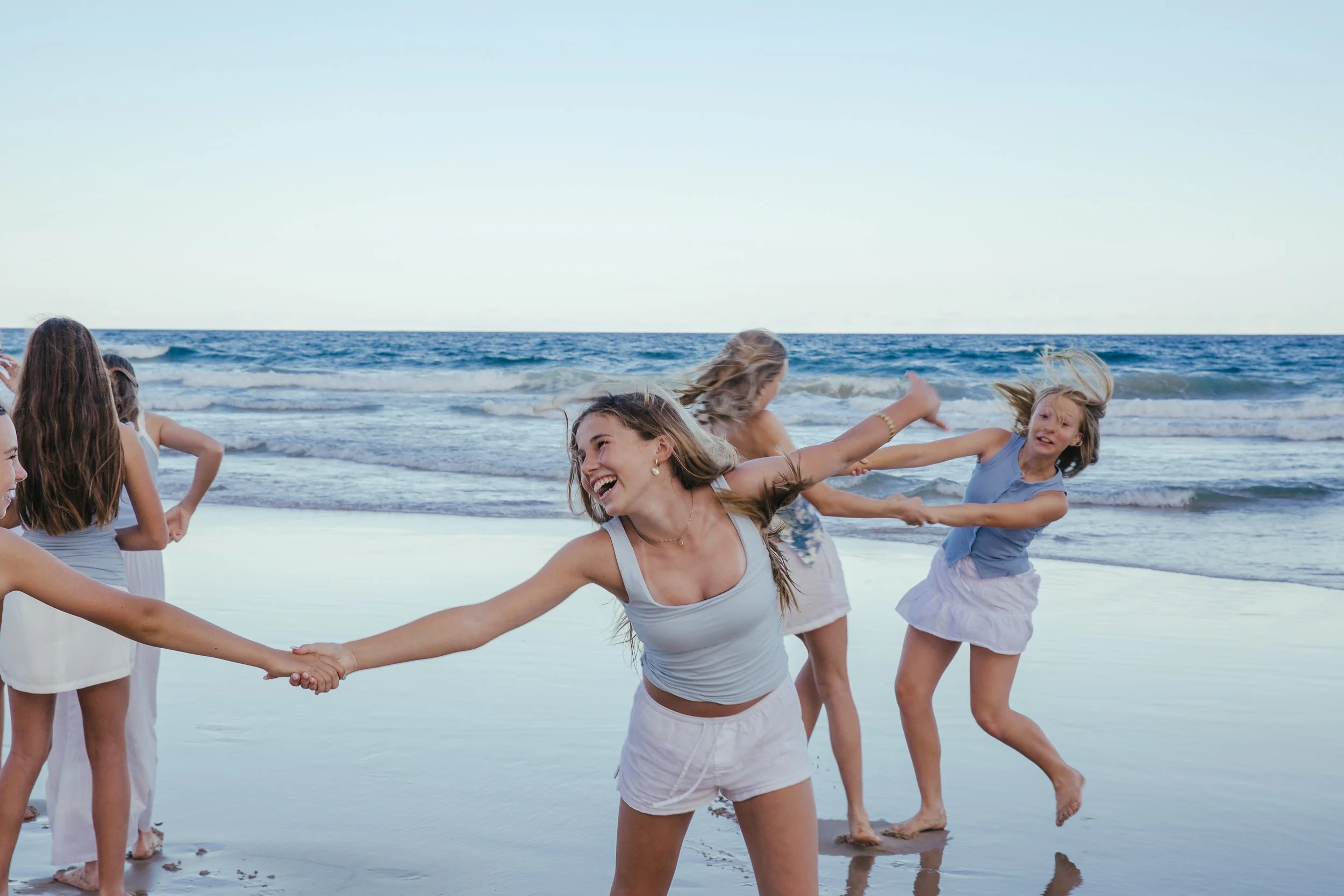 A group of women and girls holding hands and playing on a beach with waves in the background.