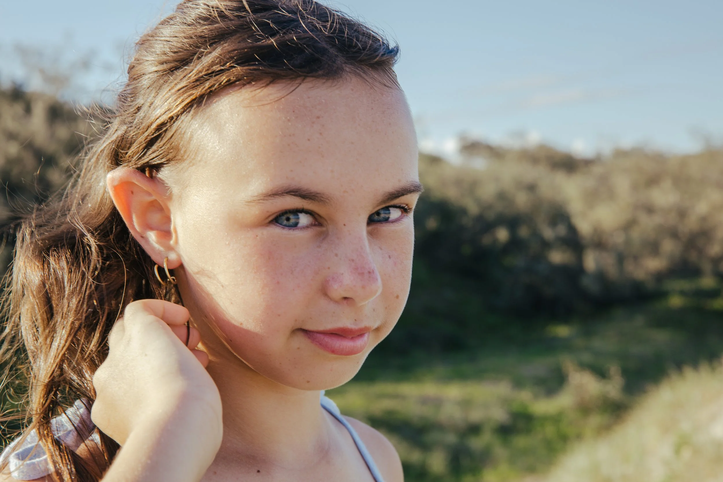 Close-up of a young girl with blue eyes, light freckles, and wet hair, standing outdoors in a sunny, natural setting.