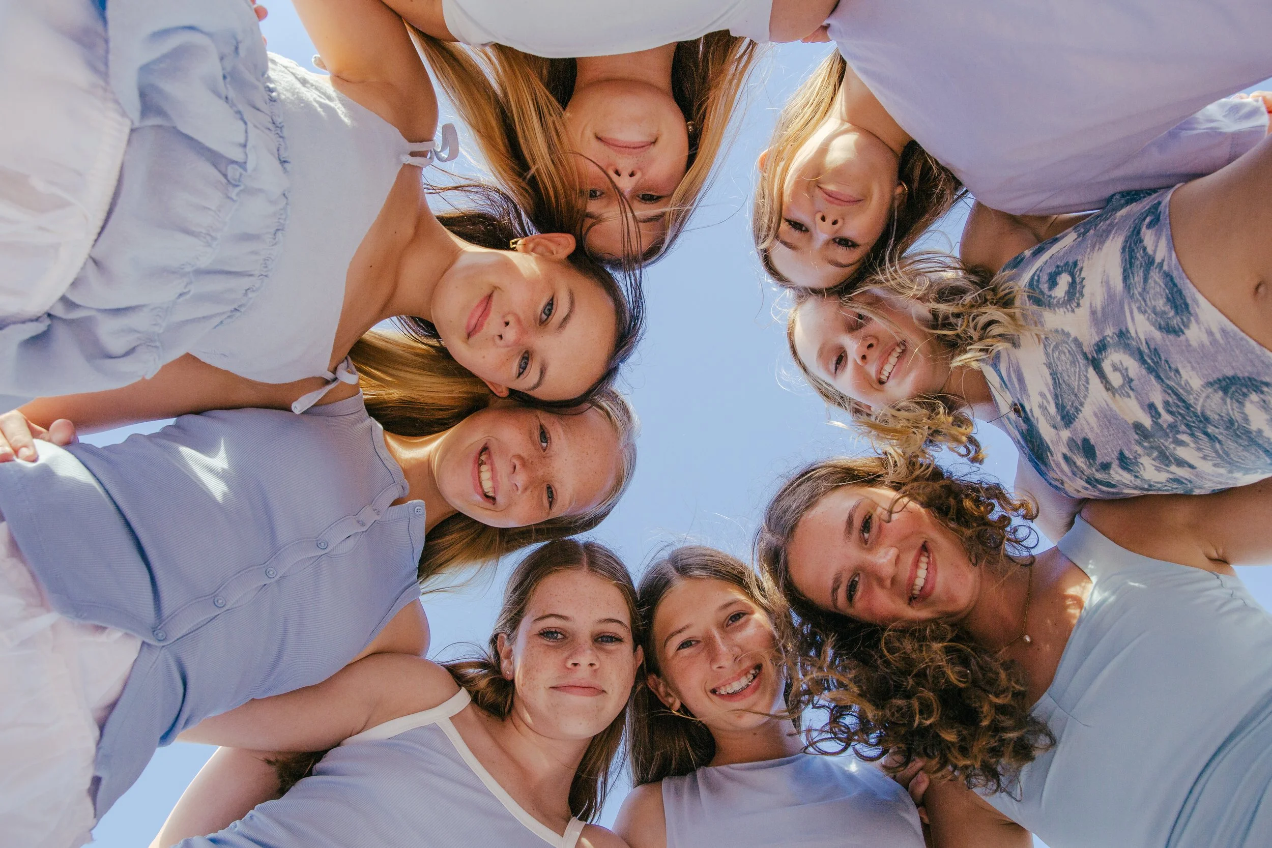 A group of young women and girls looking down into the camera, smiling, outdoors on a sunny day, with a clear blue sky.