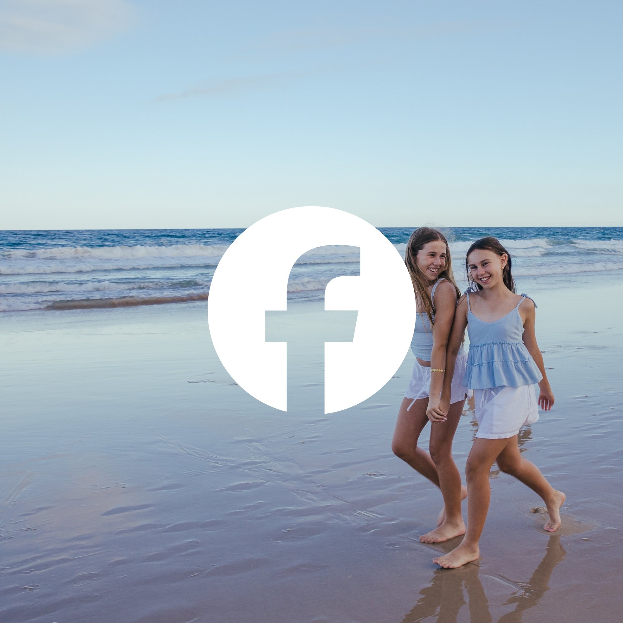 Two young women walking on the beach at sunset with the ocean in the background, smiling and looking at each other.