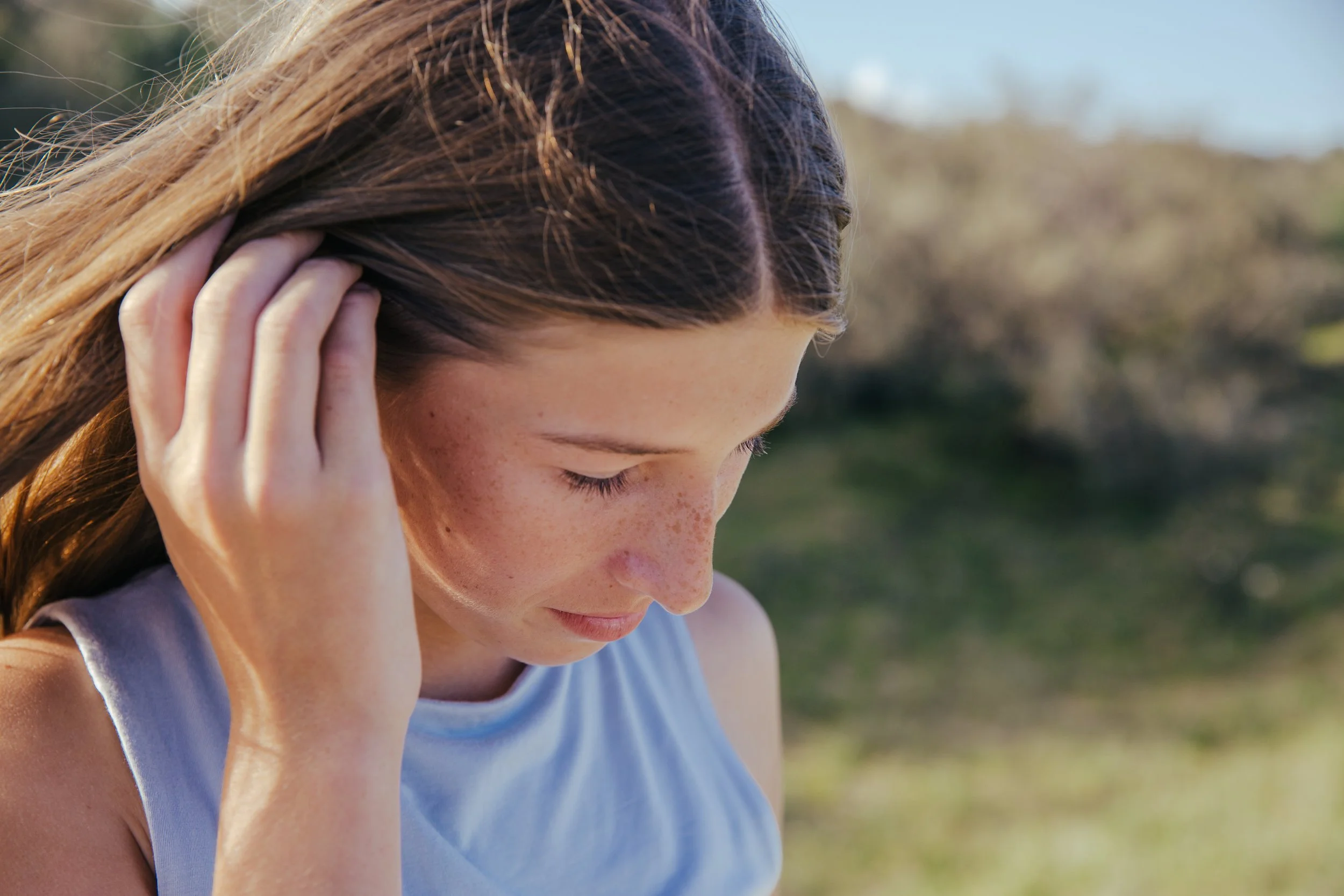 A young woman with long brown hair, wearing a light blue sleeveless top, outdoors on a sunny day, looking down with her hand touching her hair.