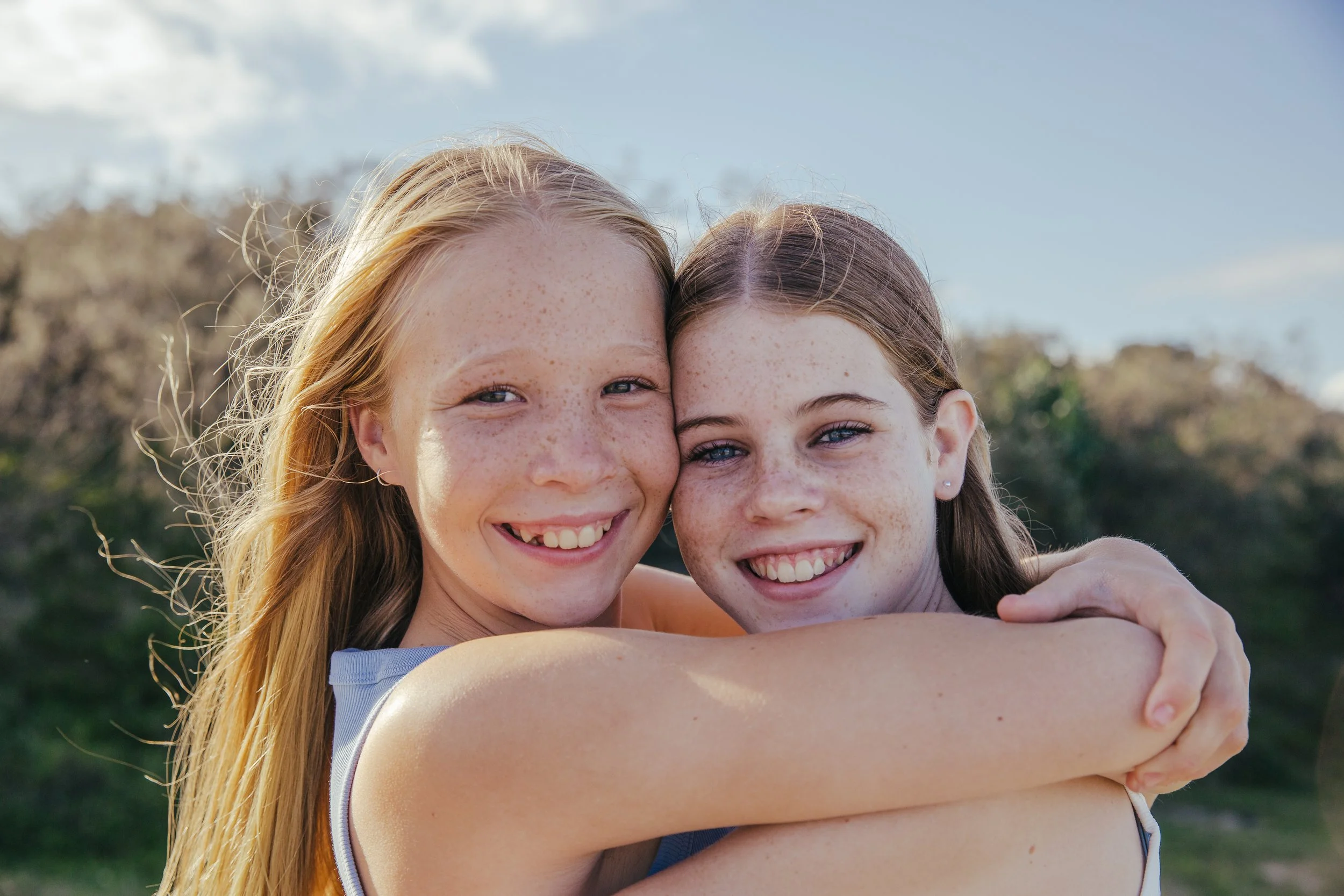 Two young women with freckles hugging outdoors under a blue sky.