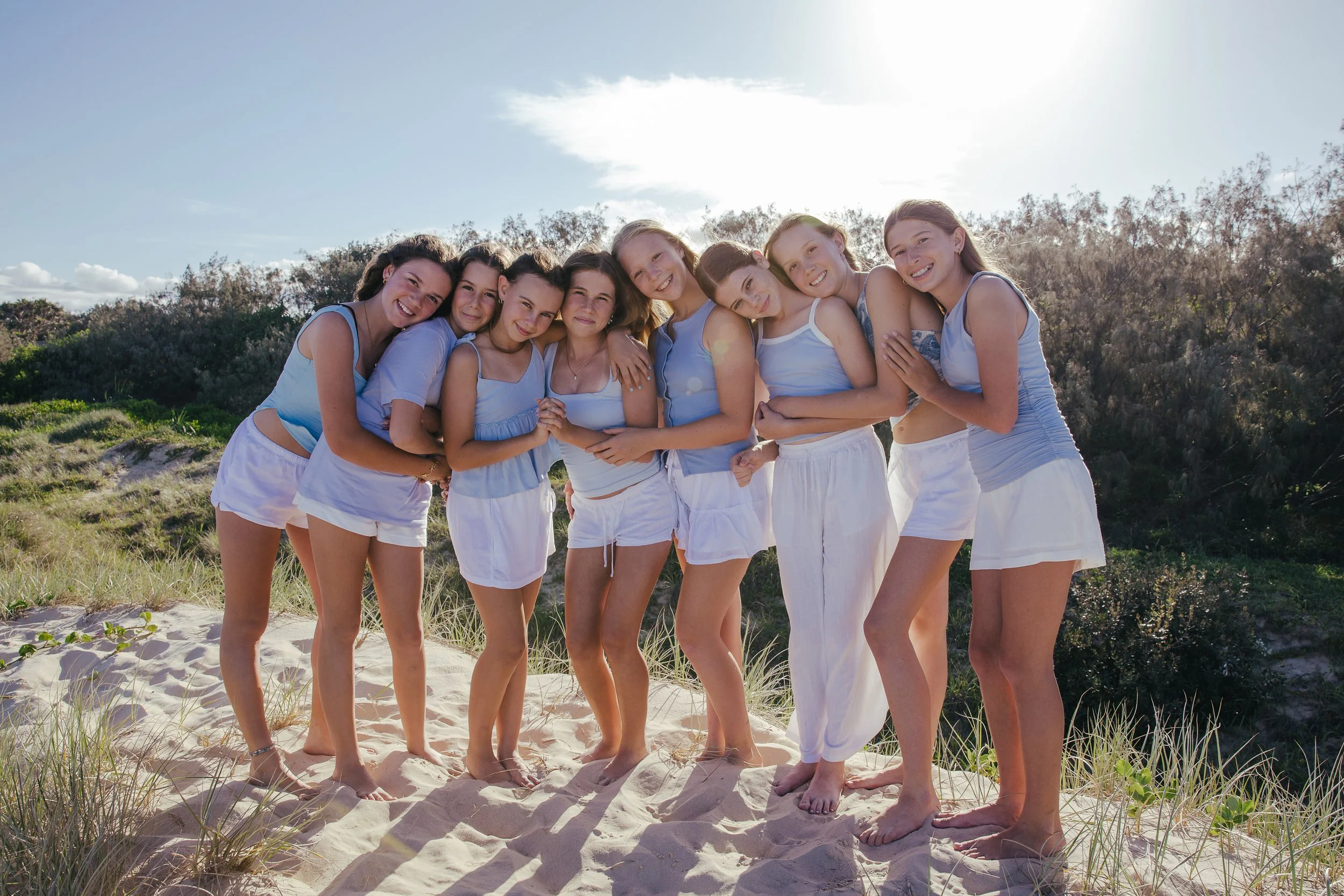 A group of eight young women standing on a sandy beach, smiling with their arms around each other, dressed in light blue and white summer clothing, with greenery and blue sky in the background.