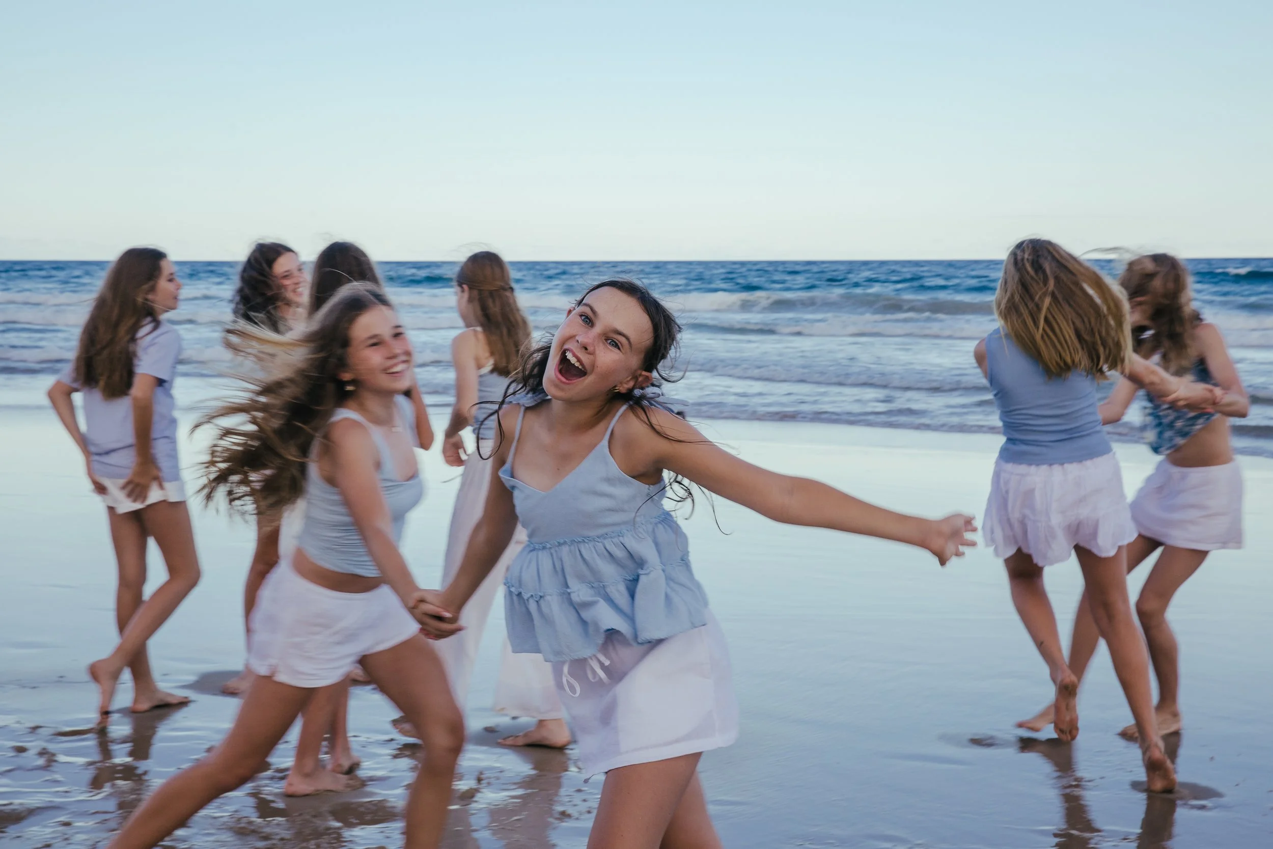 Group of young girls playing and having fun on the beach, holding hands and dancing near the shoreline with the ocean in the background.
