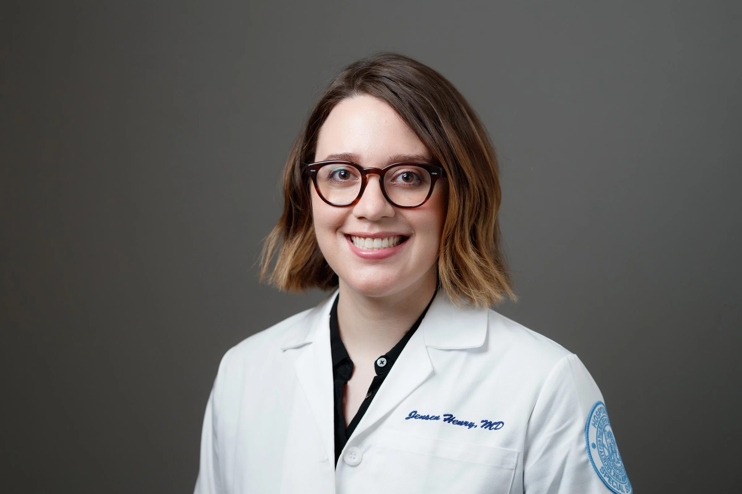 A smiling woman with short brown hair, glasses, wearing a white doctor's coat with embroidered name, standing against a neutral gray background.