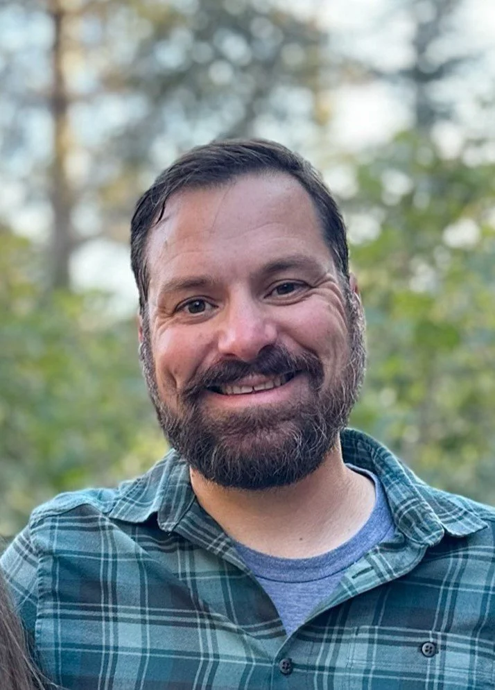 A smiling man with a beard and short dark hair, wearing a green plaid shirt and a blue t-shirt, standing outdoors with trees and sunlight in the background.