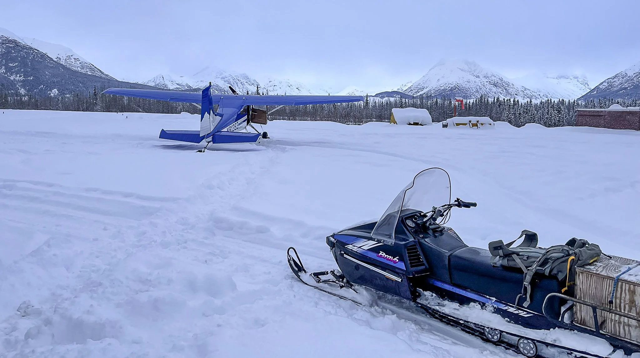 A snow-covered landscape with a blue airplane in the background and a black snowmobile in the foreground carrying luggage, mountains and trees in the distance.