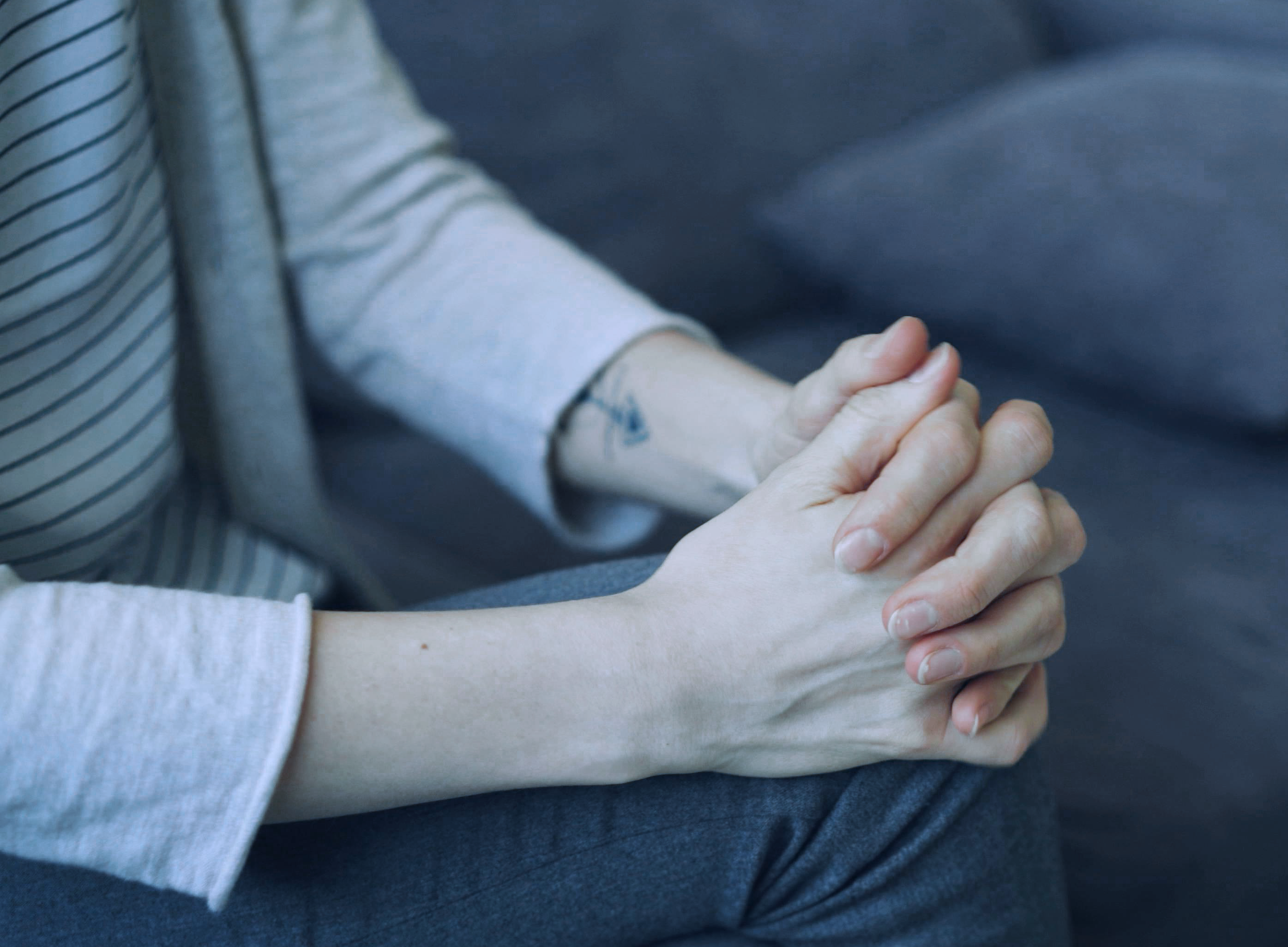 A person with light skin sitting with hands clasped on their lap. They are wearing a light gray, long-sleeved shirt with thin black stripes and dark gray pants.