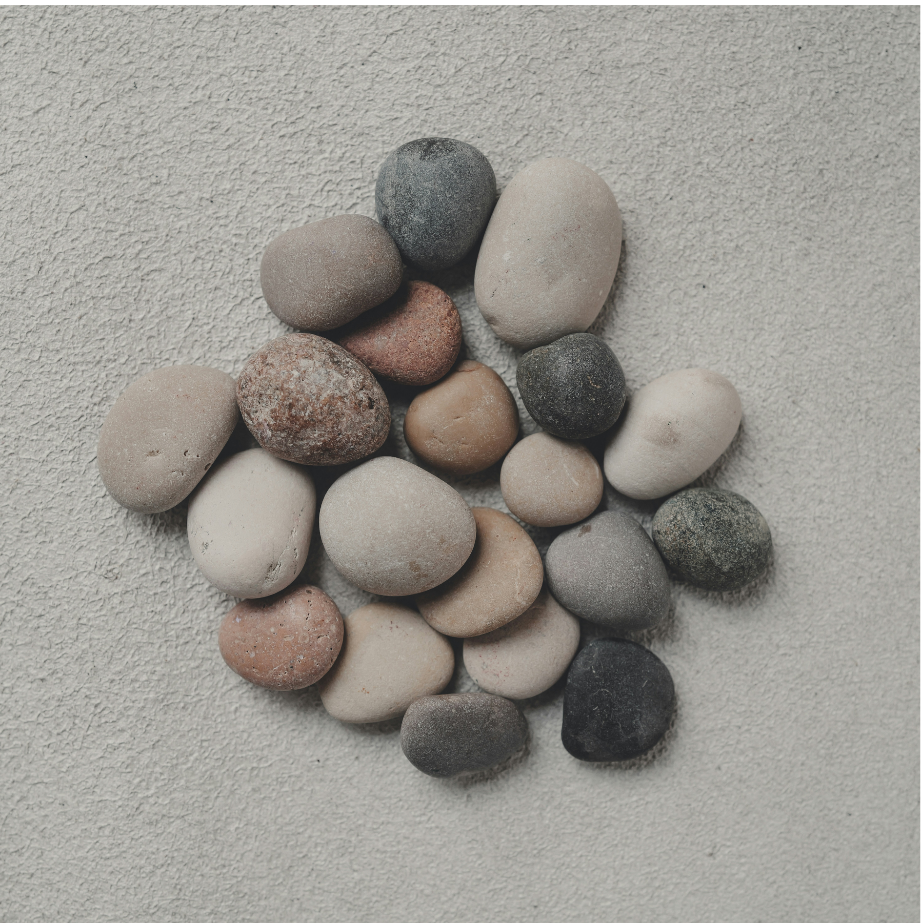 A collection of smooth, rounded stones of various colors including gray, black, white, beige, and reddish-brown, arranged on a textured light gray surface.