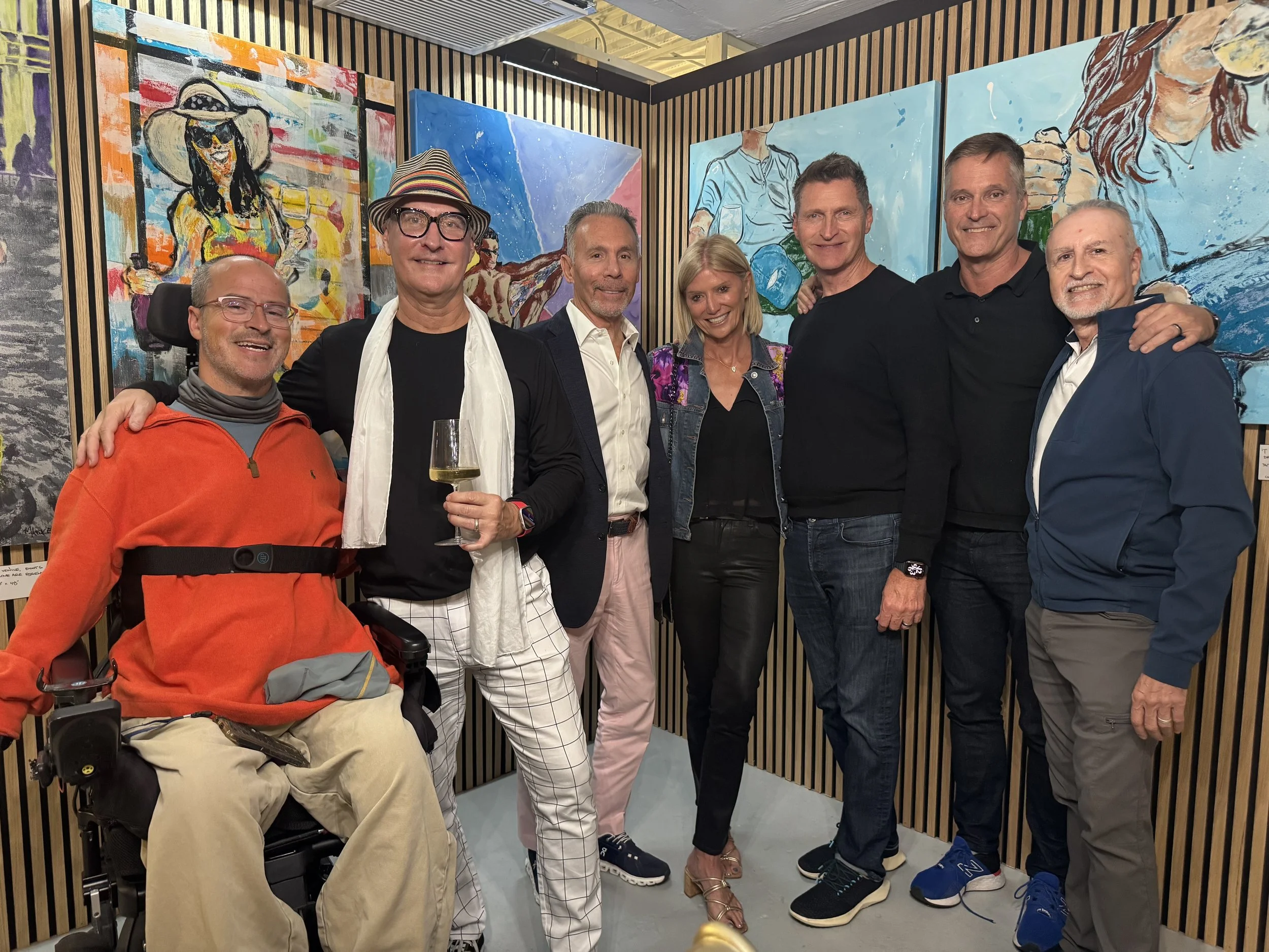 Group of seven people standing together in front of colorful paintings at an art gallery, smiling and posing for the photo.