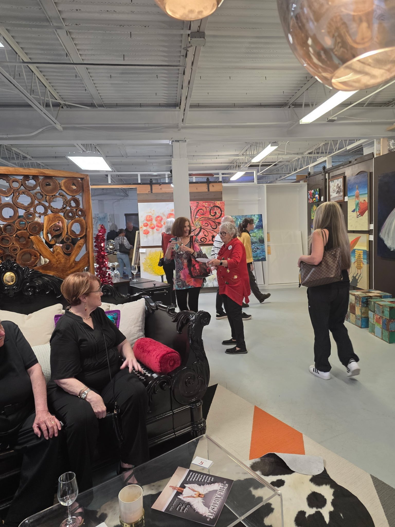 People browsing and conversing at an art gallery with various colorful paintings on the walls, an ornate black sofa, and a cowhide rug.