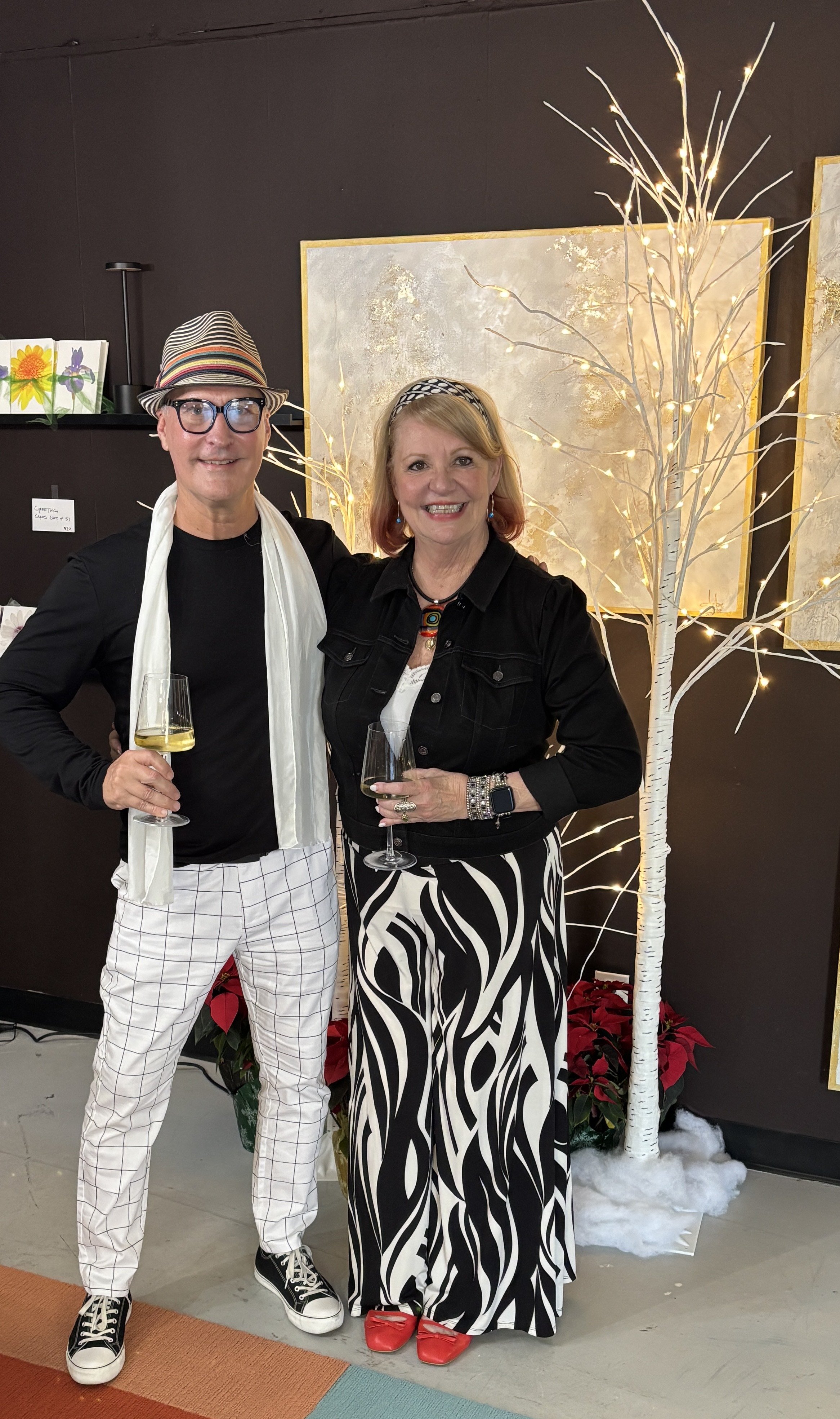 A man and woman standing together, smiling, holding glasses of white wine at a festive event, with decorative lights and a winter-themed scene behind them.