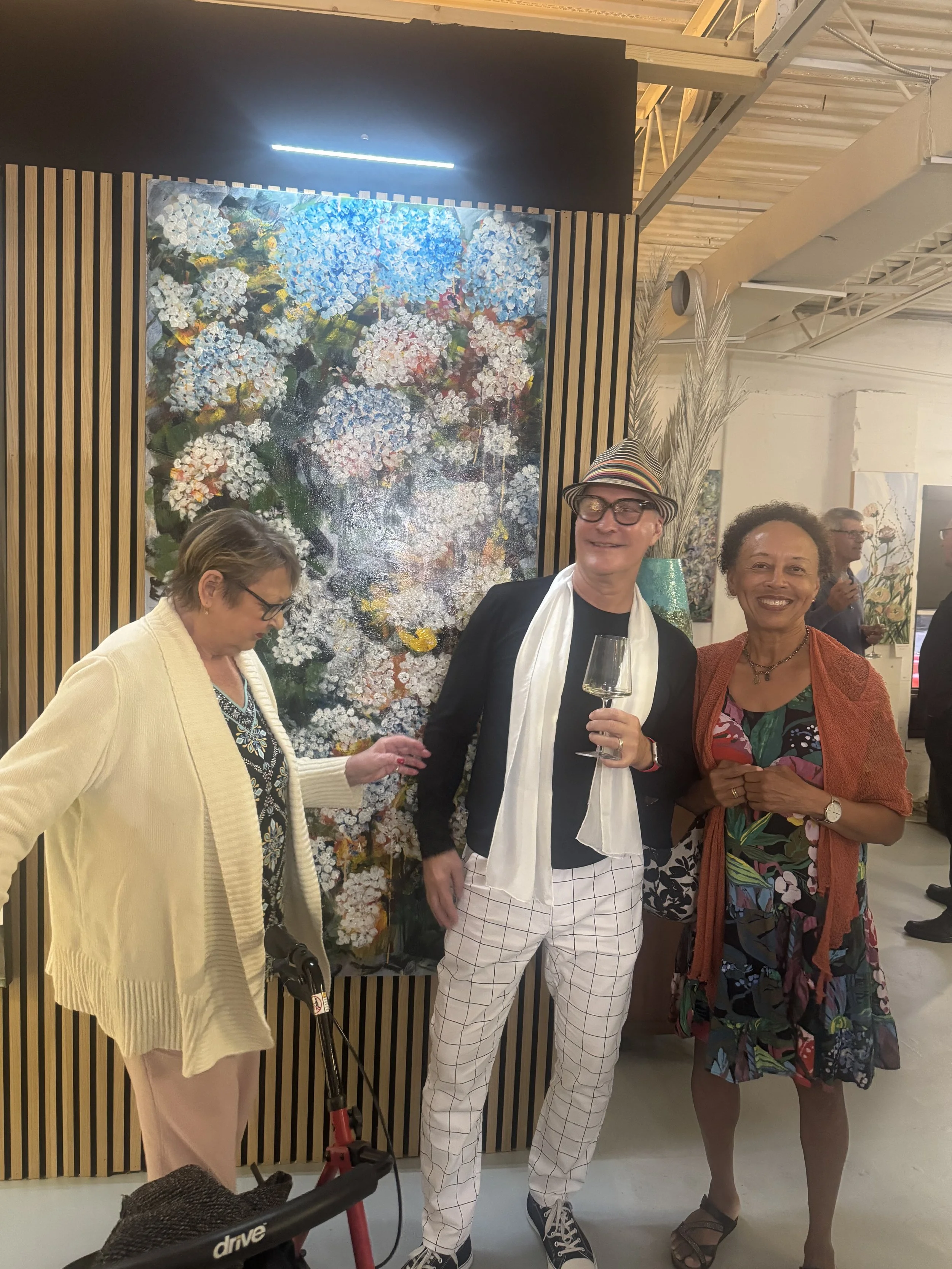 Three people at an art gallery, with a painting of flowers behind them. The man in the middle wears glasses, a striped hat, a white scarf, checkered pants, and is holding a glass of wine. The woman on the right wears a colorful dress with a rust-colored shawl, and the woman on the left wears a beige sweater and glasses.