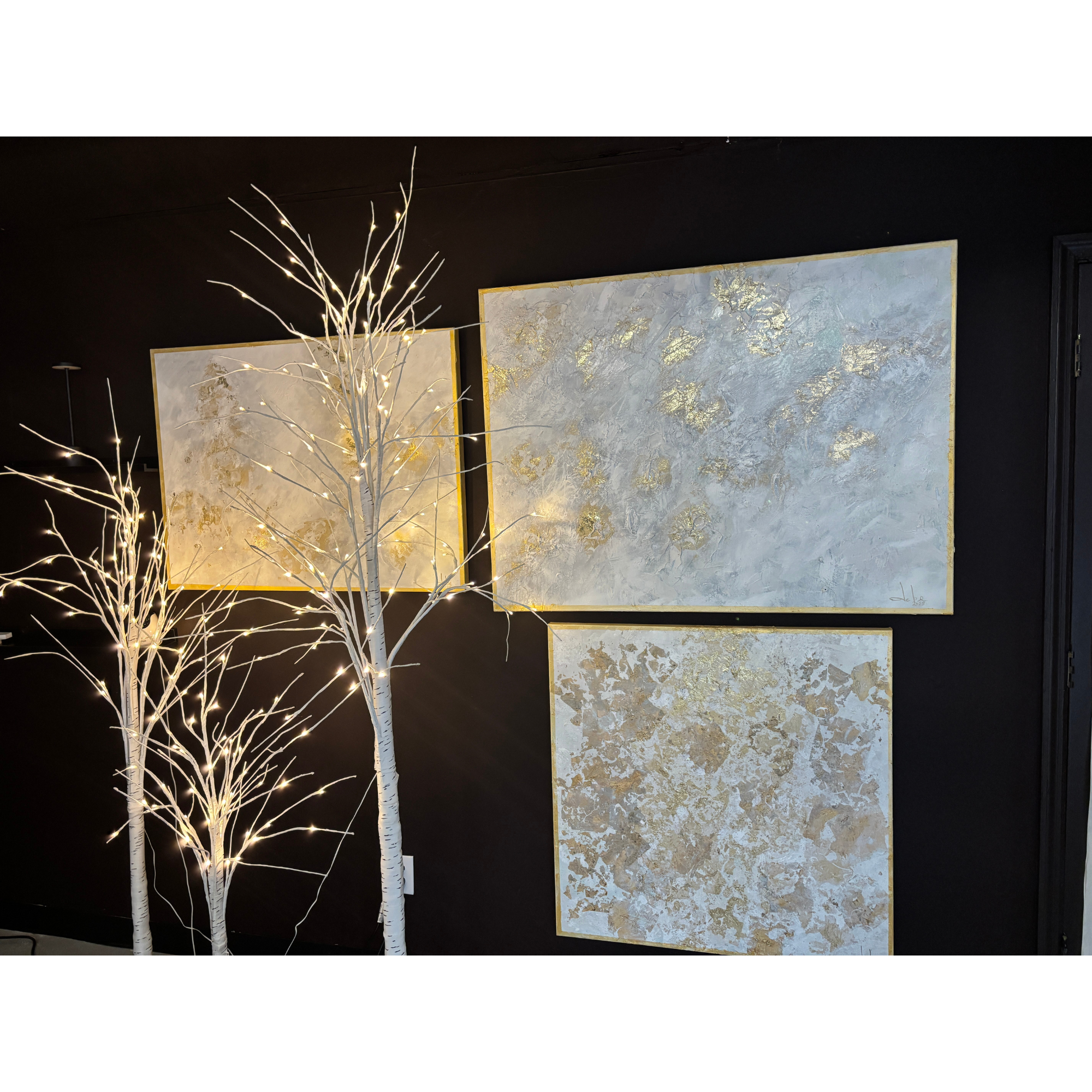 Decorative wall art with three textured abstract paintings in white, cream, and gold tones, illuminated by white string lights on leafless white branches against a black wall.