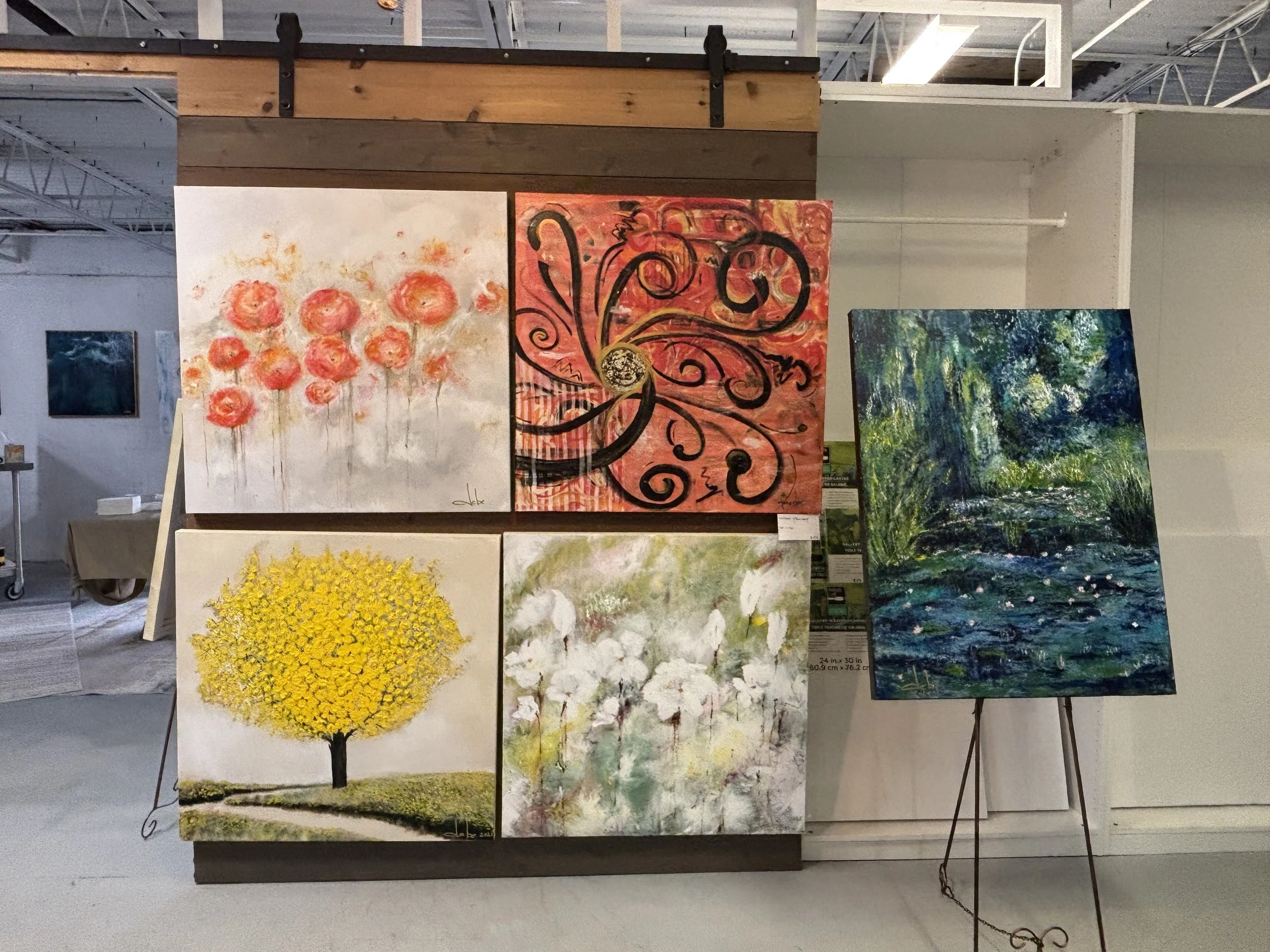 Display of five colorful paintings at an art gallery. The top left painting depicts pink flowers on a white background. The top center painting has a swirling black and red abstract design. The bottom left painting shows a bright yellow tree on a green hill. The bottom center painting features white flowers with green and yellow accents. The right painting is an impressionistic landscape of a forest with a creek, rendered in blue, green, and white tones.