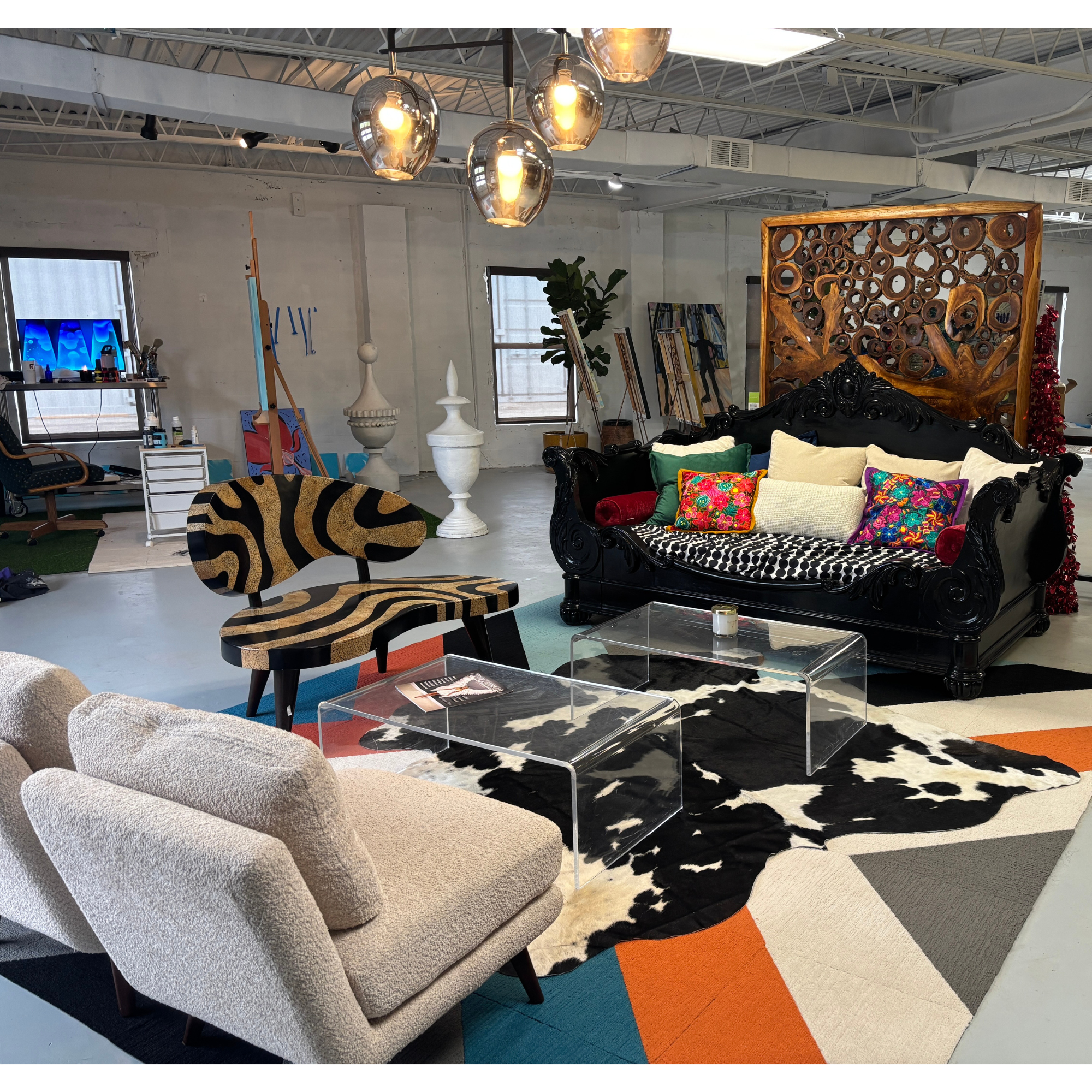 Interior of a stylish living room with a beige armchair, black ornate sofa with colorful pillows, zebra-patterned rug, clear acrylic coffee tables, and various decorative items including a large wooden art piece and a wall background of windows and industrial ceiling.