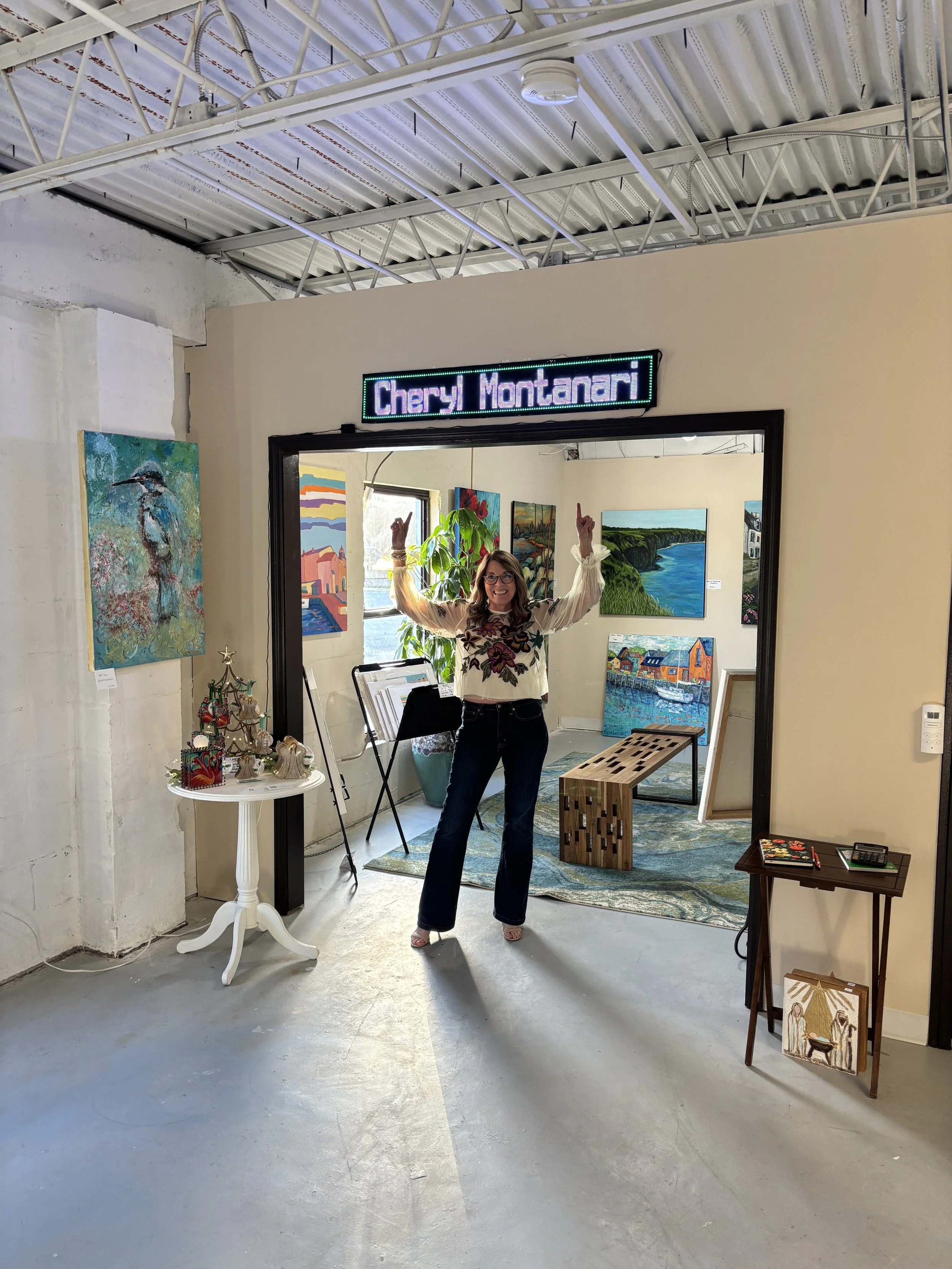 Woman standing inside art gallery, smiling, raising her arms, with paintings on the walls behind her.