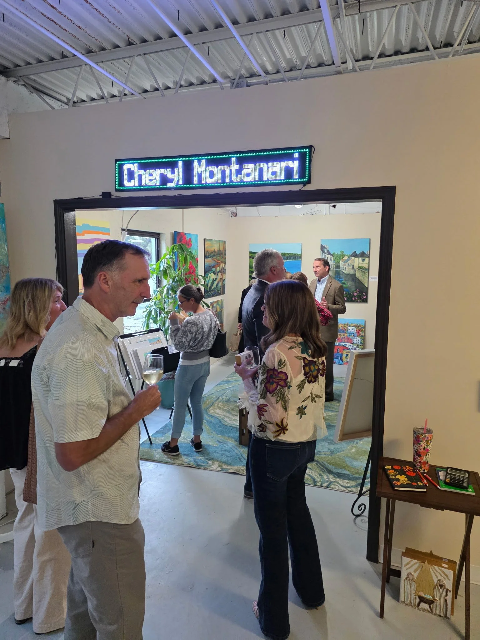 People attending an art gallery opening, with paintings on the walls, a table with art supplies, and a sign with the name 'Cheryl Montanari' above the entrance.